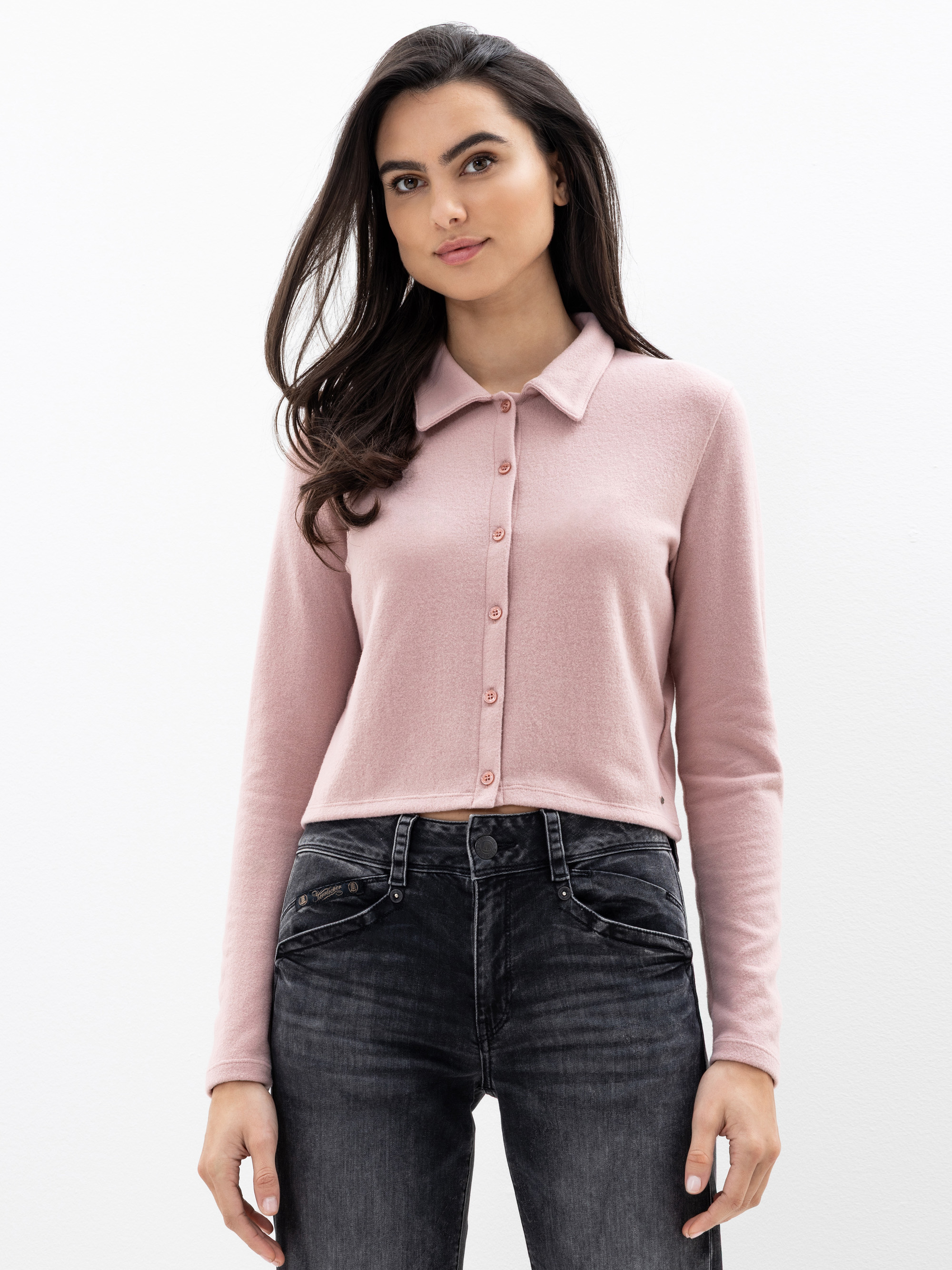 Eine Frau mit langen dunklen Haaren trägt eine hellrosa Strickjacke mit Kragen und dunkle Jeans mit hoher Taille vor einem schlichten weißen Hintergrund.