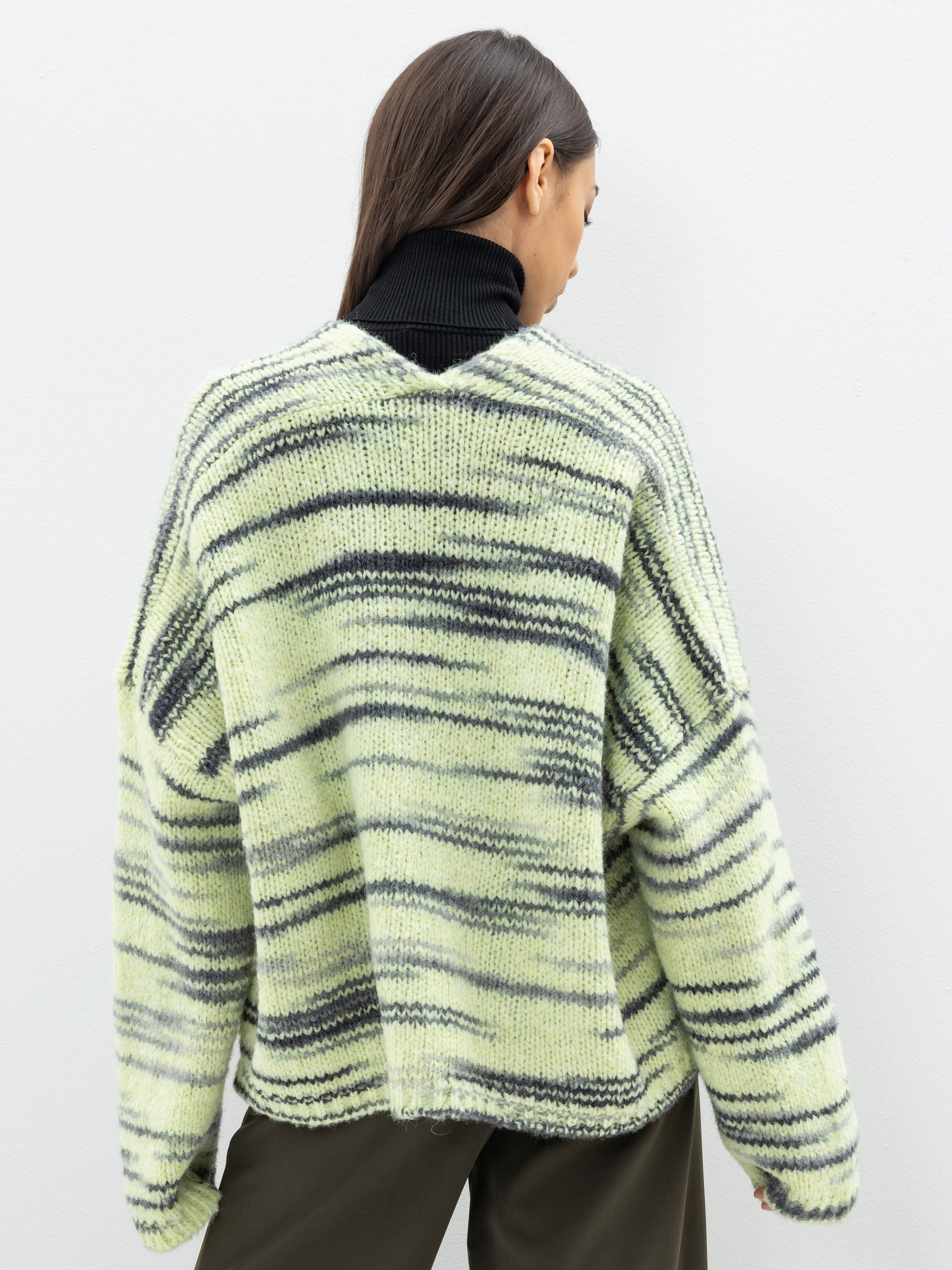 Eine Person mit langem, glattem braunem Haar wird von hinten gezeigt, die einen lockeren, grün-schwarz gestreiften Strickpullover über einem schwarzen Rollkragenpullover und eine dunkle Hose trägt und vor einer schlichten hellen Wand steht.