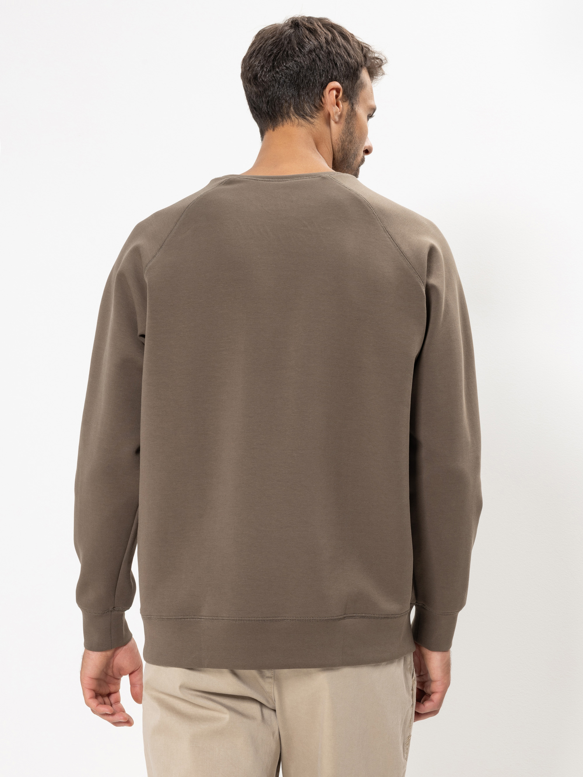 Ein Mann steht mit einem hellbraunen Sweatshirt und einer beigen Hose vor einem einfarbig weißen Hintergrund und schaut von der Kamera weg.