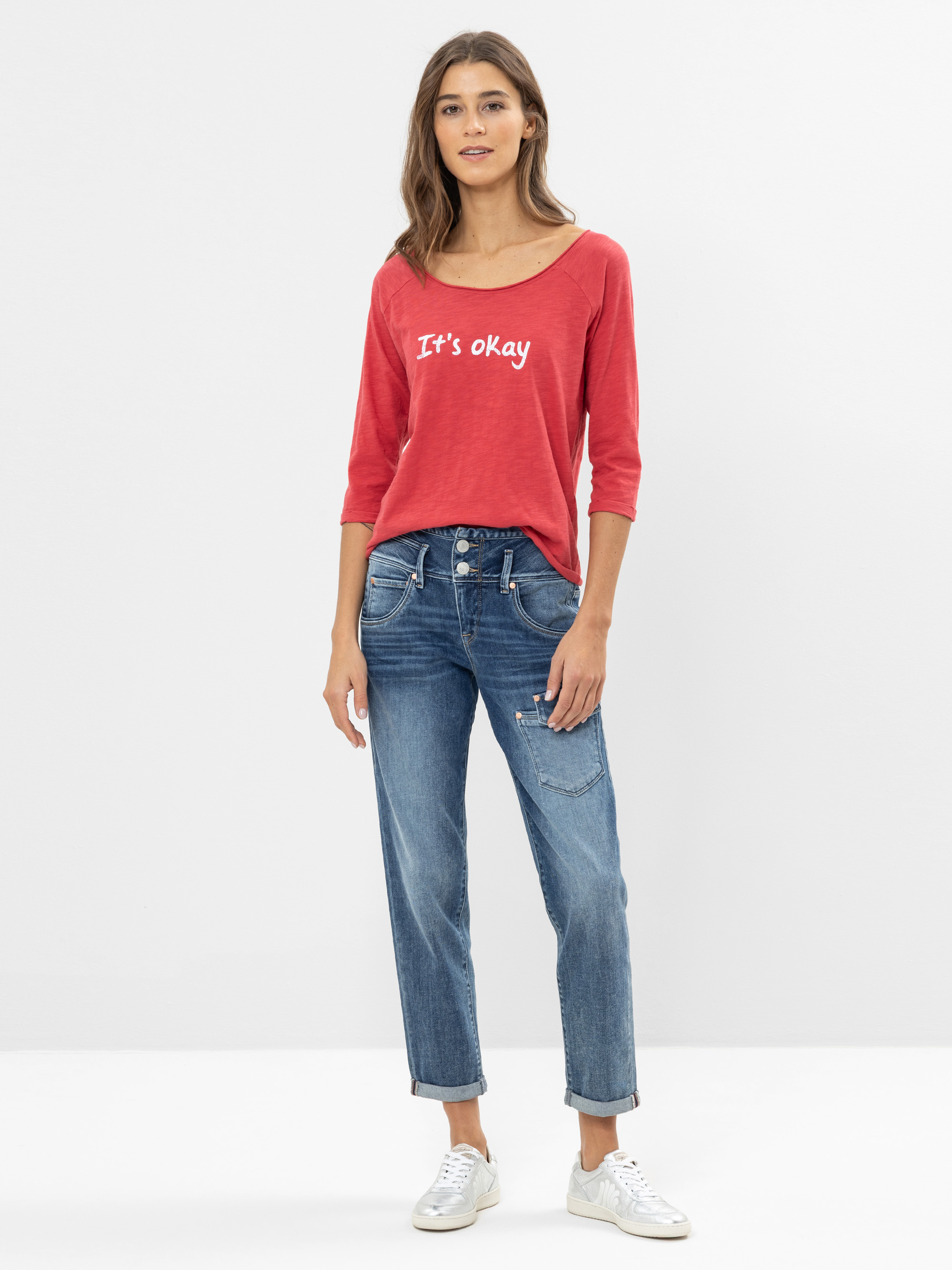 Eine Frau steht vor einem schlichten weißen Hintergrund und trägt ein rotes langärmeliges Hemd mit der Aufschrift Its okay, blaue Jeans und weiße Turnschuhe. Sie hat langes braunes Haar und einen entspannten Gesichtsausdruck.