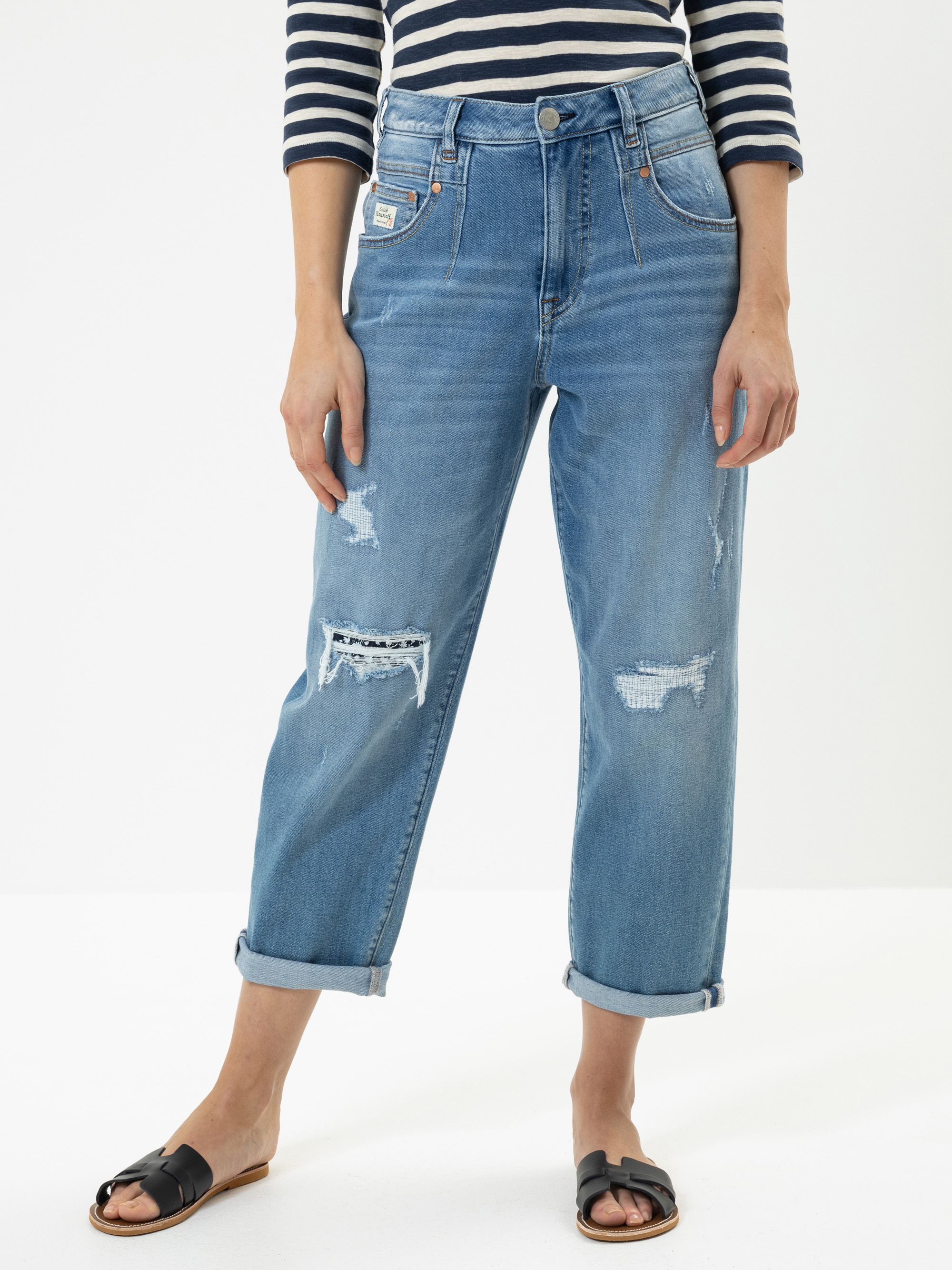 Eine Person, die ein marineblau-weiß gestreiftes langärmeliges Oberteil, hellblaue Jeans mit aufgerollten Bündchen und schwarze Slide-Sandalen trägt, steht vor einem schlichten weißen Hintergrund.