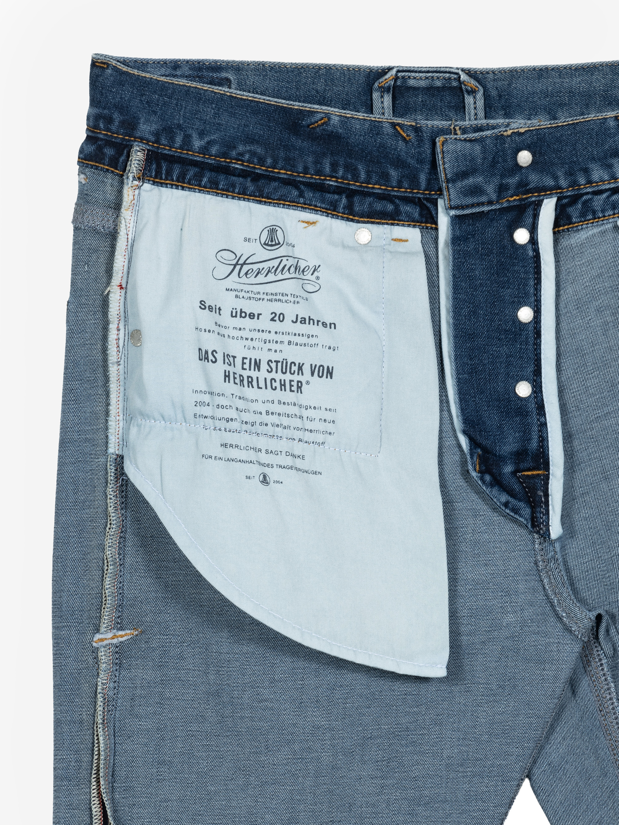 Eine Nahaufnahme einer blauen Jeans mit geöffnetem Hosenschlitz, die das Innenfutter der Taschen zeigt, auf dem ein deutscher Text und der Markenname Herrlicher aufgedruckt sind. Der Hosenschlitz mit Knöpfen und die Denim-Struktur sind deutlich zu erken