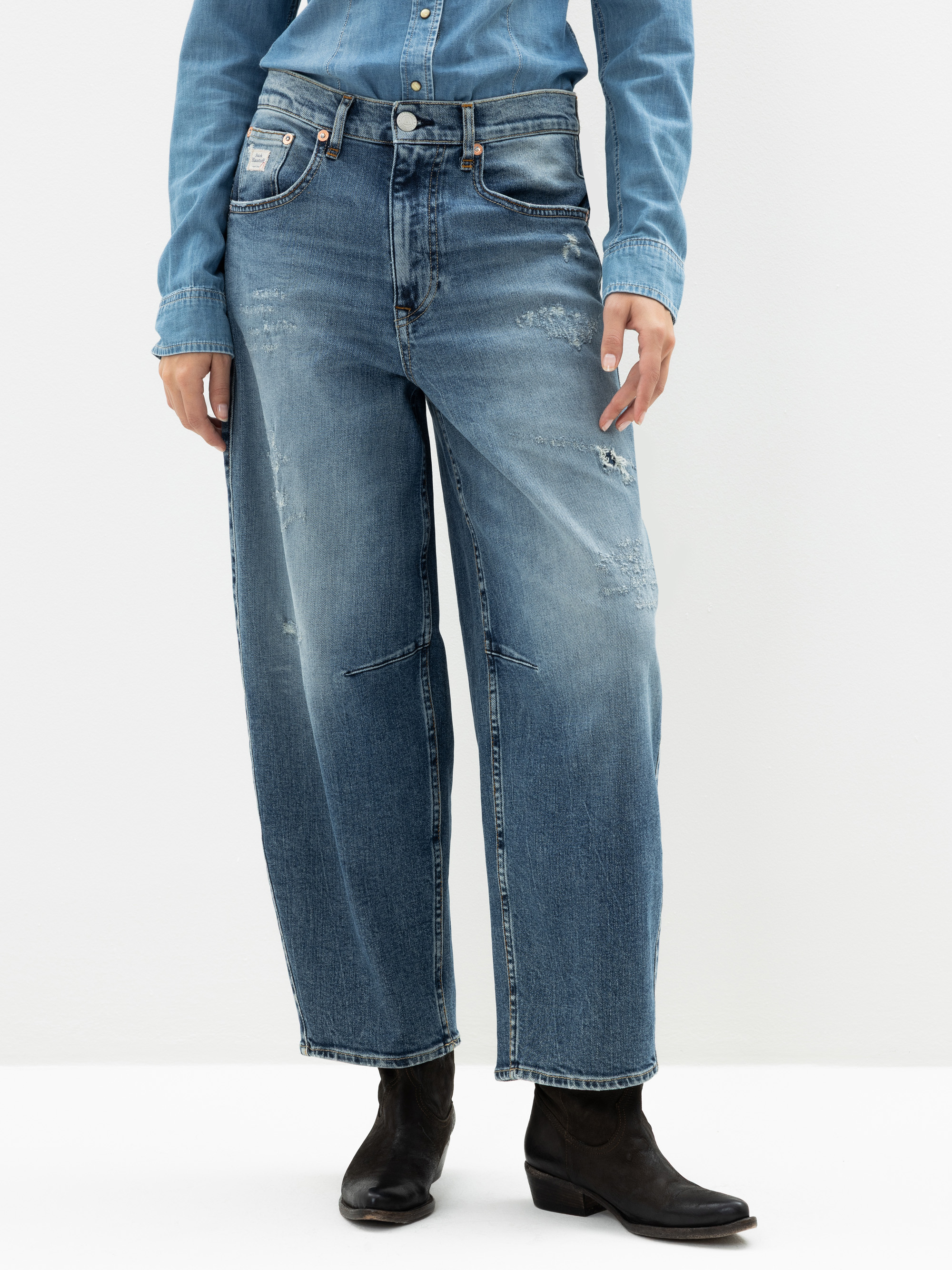 Eine Person, die locker sitzende, leicht abgenutzte Bluejeans, ein in die Hose gestecktes Jeanshemd und schwarze Ankle Boots trägt, steht vor einem schlichten weißen Hintergrund.