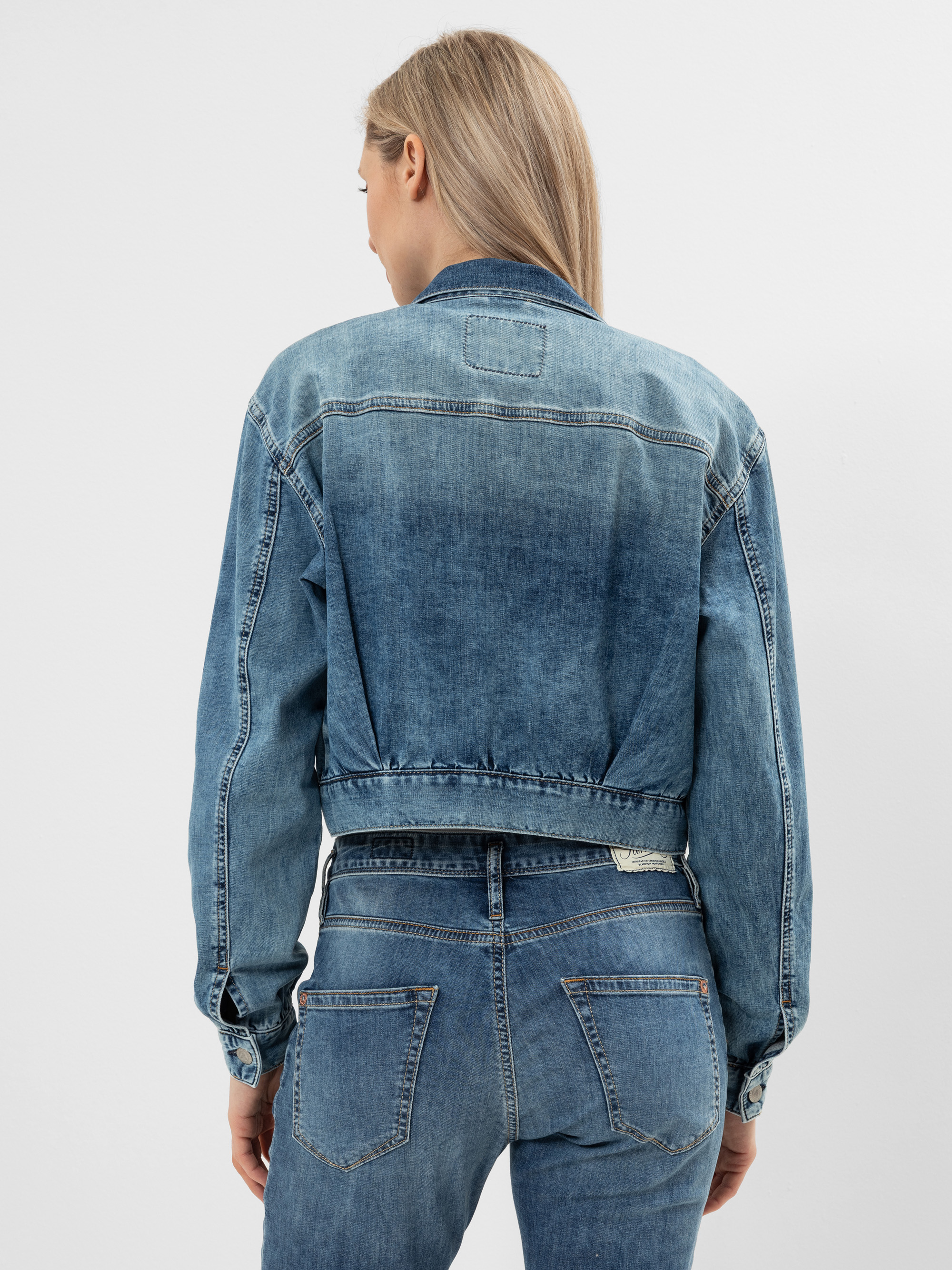 Eine Frau mit glattem, hellbraunem Haar steht mit dem Rücken zur Kamera und trägt eine blaue Jeansjacke und Jeanshosen vor einem schlichten weißen Hintergrund.