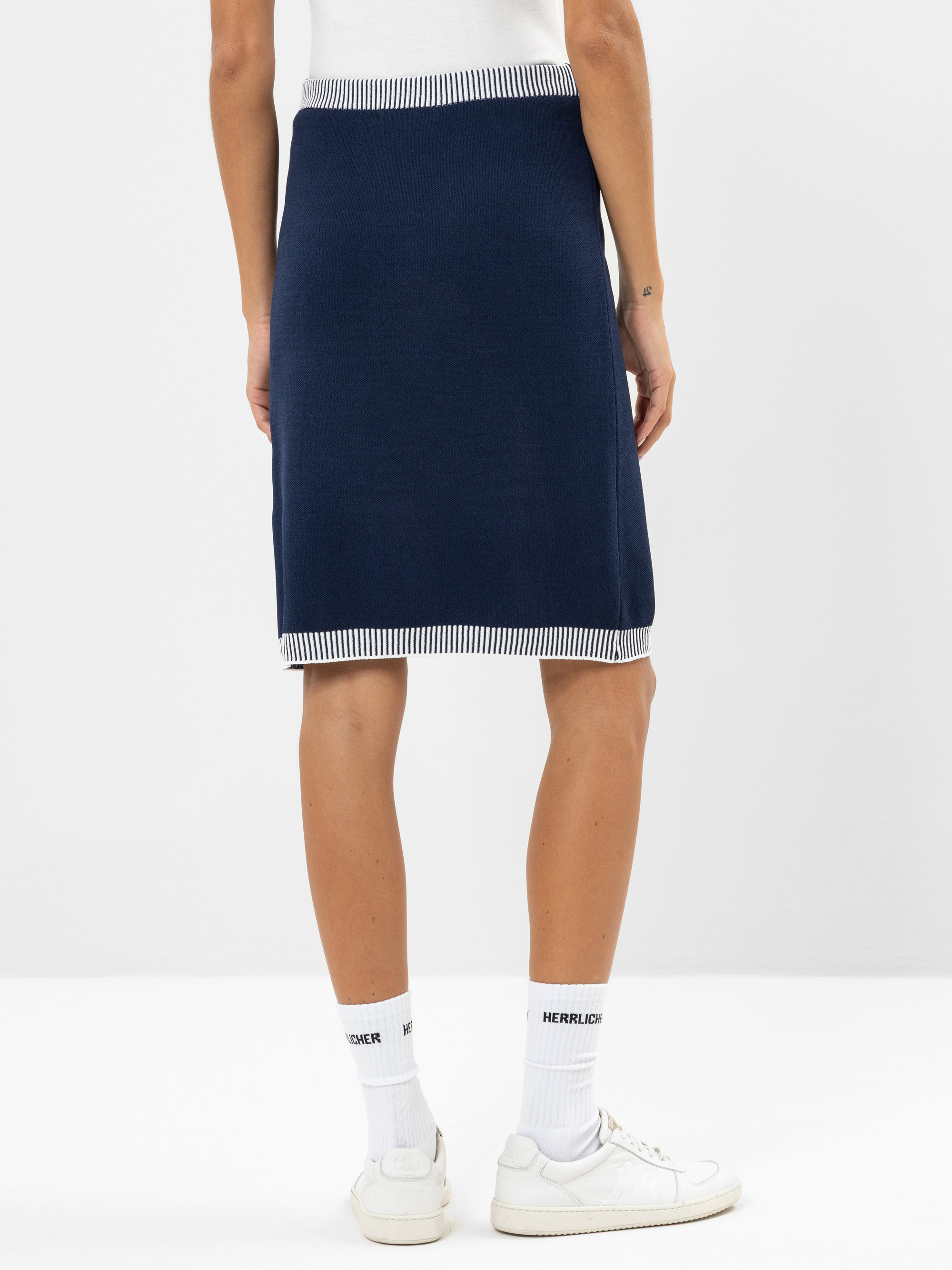 Eine Person steht mit dem Gesicht zur Wand und trägt einen marineblauen knielangen Rock mit weißem und marineblauem Streifenmuster, weiße Socken mit der Aufschrift HERZLIEBER und weiße Turnschuhe vor einem unifarbenen Hintergrund.