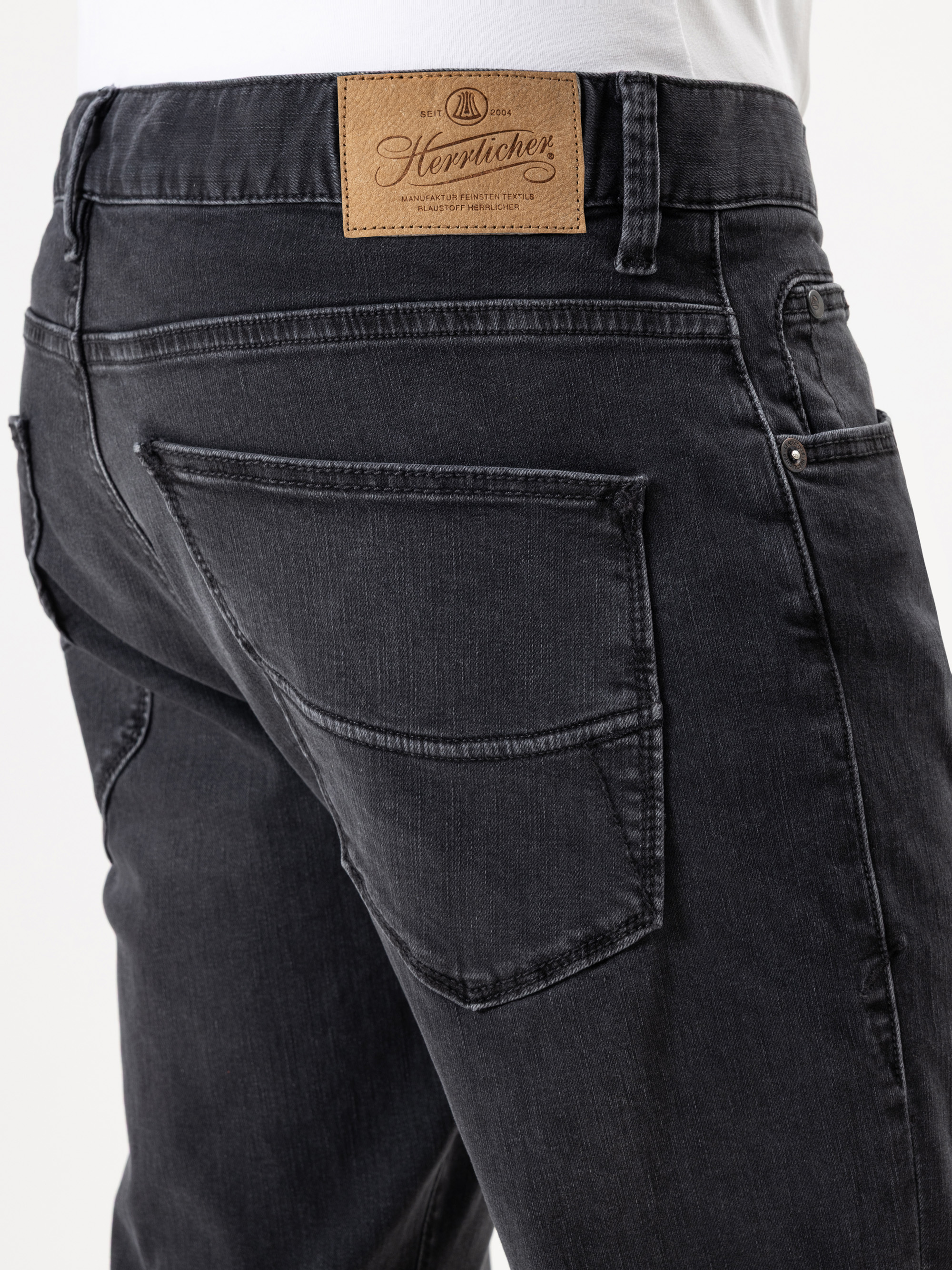 Nahaufnahme des Rückens einer Person, die eine schwarze Denim-Jeans mit großen Gesäßtaschen und einem braunen Lederaufnäher der Marke Herrlicher am Bund trägt.