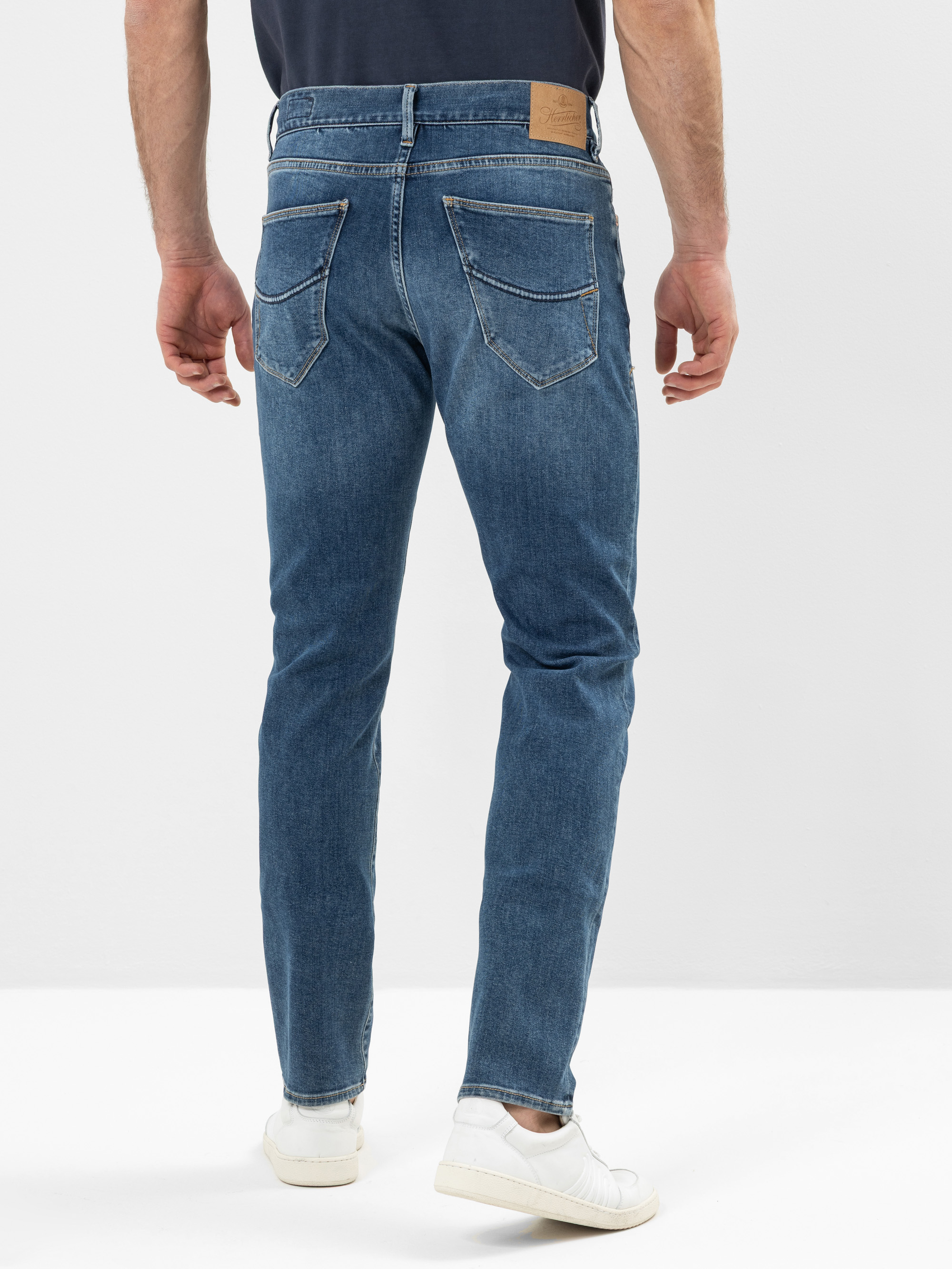 Ein Mann in blauen Jeans und weißen Turnschuhen steht mit dem Gesicht zur Kamera vor einem schlichten weißen Hintergrund. Die Jeans hat zwei Gesäßtaschen und einen braunen Aufnäher am Bund.