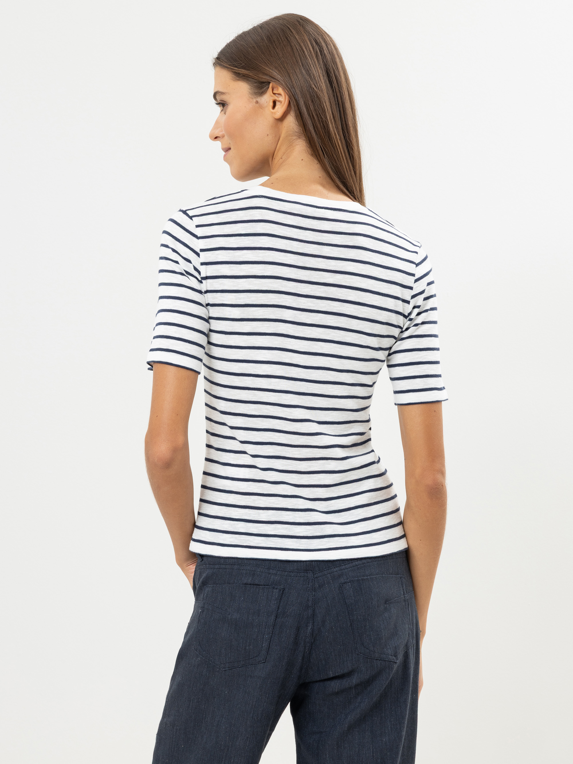 Eine Frau mit langen braunen Haaren steht mit dem Gesicht nach hinten, trägt ein weißes T-Shirt mit horizontalen marineblauen Streifen und eine dunkelblaue Hose und steht vor einem schlichten hellen Hintergrund.