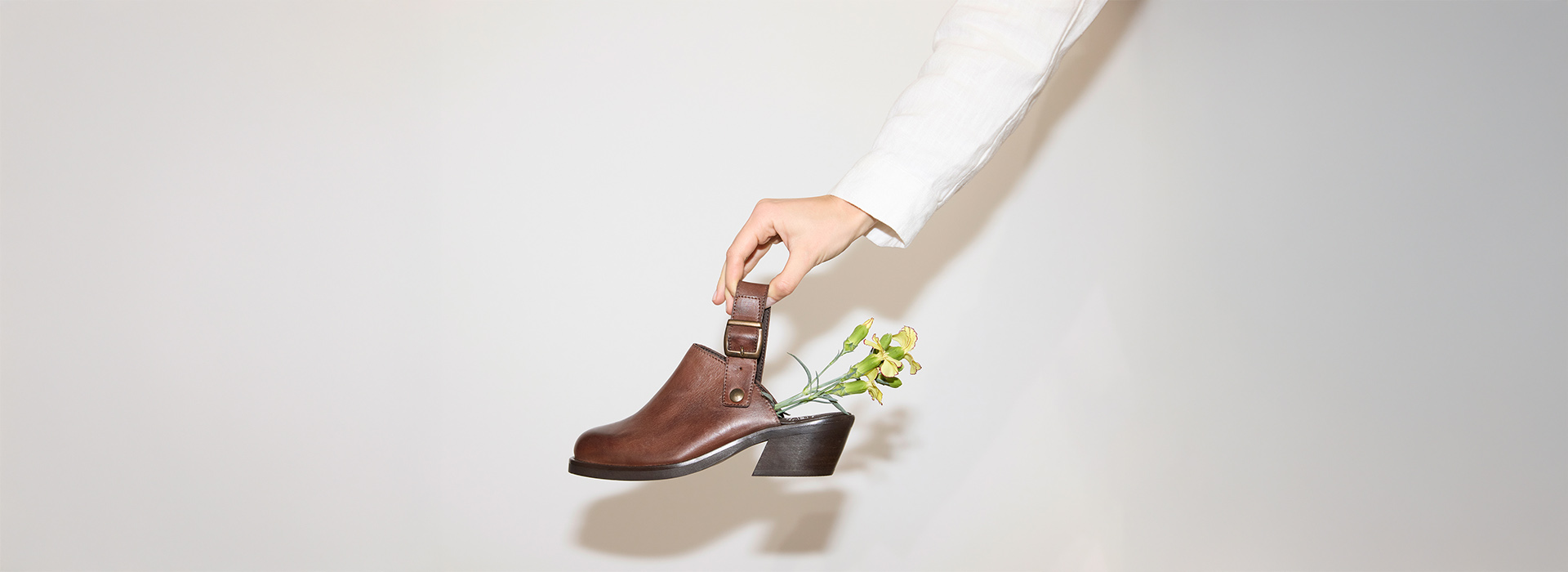 Eine Hand in einem weißen Ärmel hält einen braunen Schuh mit Absatz, in dem sich grüne Blumen befinden, vor einem schlichten hellen Hintergrund.