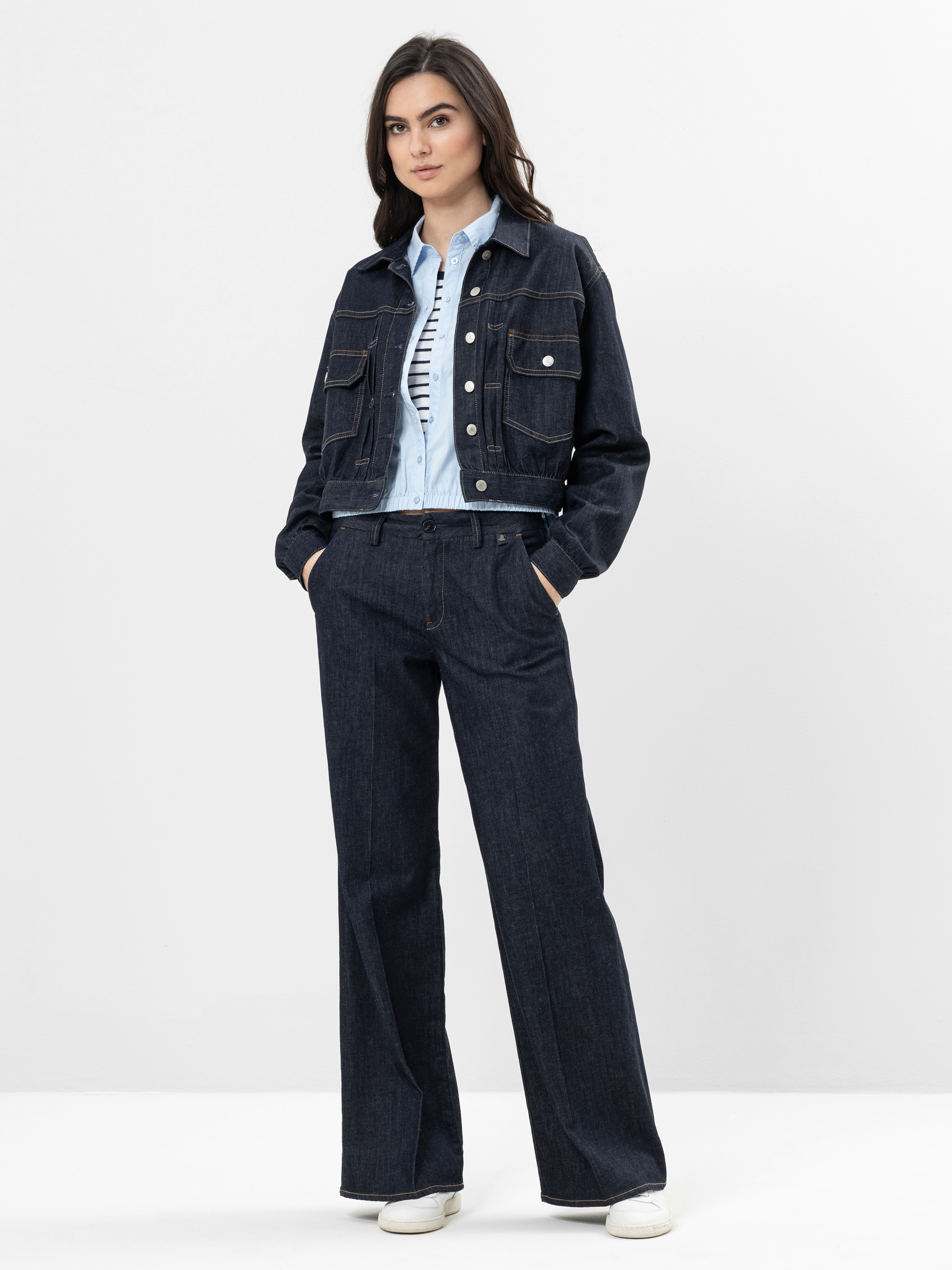 Eine Frau steht vor einem schlichten Hintergrund und trägt eine dunkle Jeansjacke über einem hellblauen Hemd, eine dunkle Jeanshose mit weitem Bein und weiße Turnschuhe. Sie hat langes dunkles Haar und einen neutralen Gesichtsausdruck.