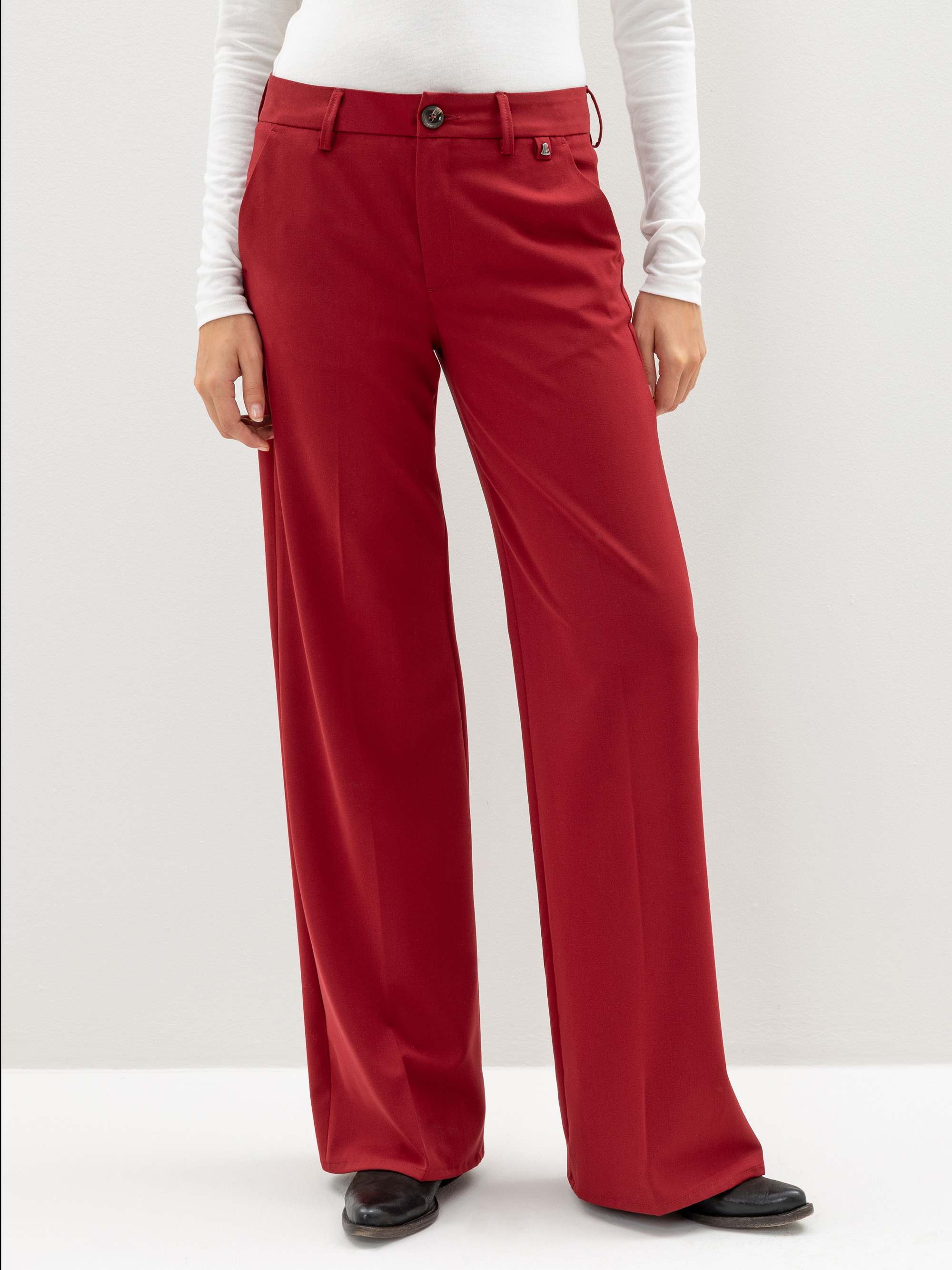 Eine Person, die eine rote Hose mit weitem Bein und hoher Taille, ein weißes, langärmeliges Oberteil und schwarze Schuhe trägt, steht vor einem einfarbigen, hellen Hintergrund. Das Bild ist an den Schultern abgeschnitten.