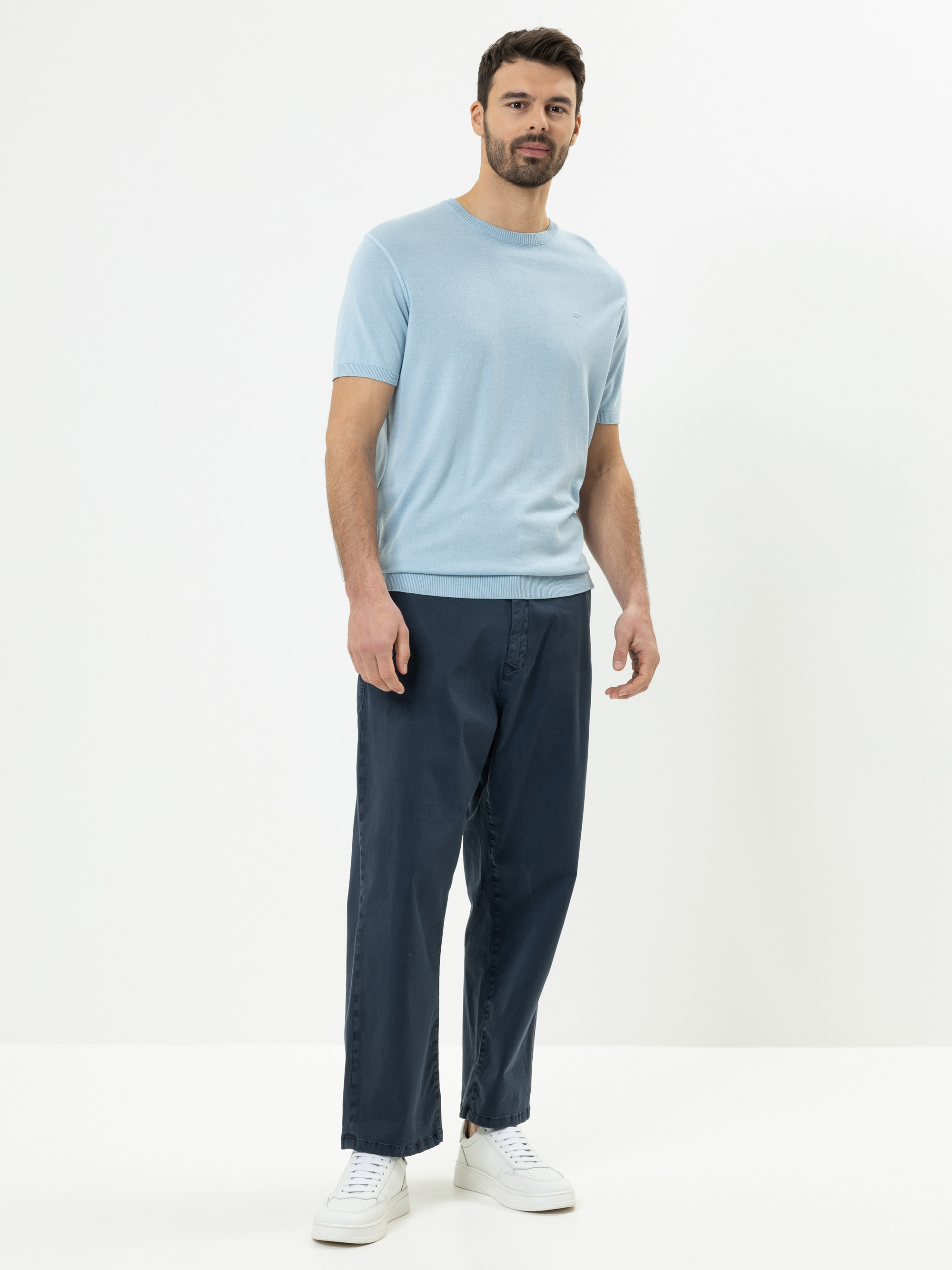 Ein Mann steht vor einem schlichten weißen Hintergrund und trägt ein hellblaues kurzärmeliges Hemd, eine dunkelblaue Hose und weiße Turnschuhe.