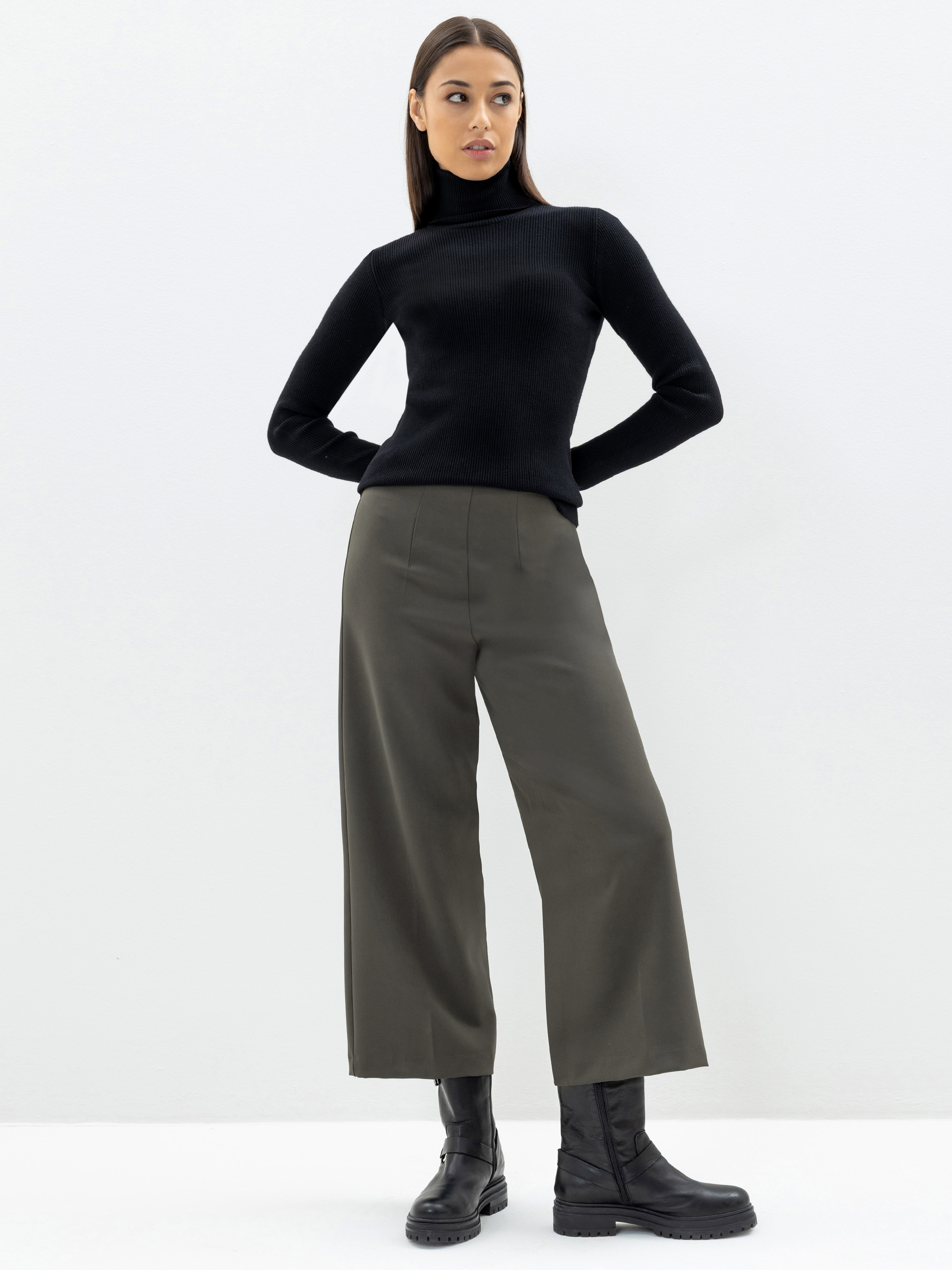 Eine Frau steht vor einem weißen Hintergrund und trägt einen schwarzen Rollkragenpullover, eine olivgrüne Hose mit weitem Bein und schwarze Stiefel. Sie hat glattes braunes Haar und einen neutralen Gesichtsausdruck, die Hände sind in die Hüften geste