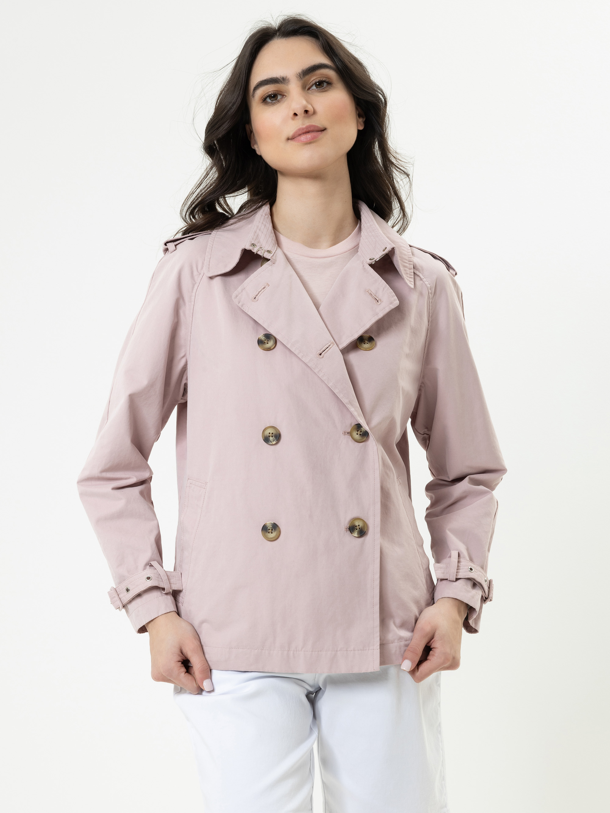 Eine Frau mit langen dunklen Haaren trägt einen hellrosa zweireihigen Trenchcoat über einem passenden Oberteil und einer weißen Hose und steht vor einem schlichten weißen Hintergrund.