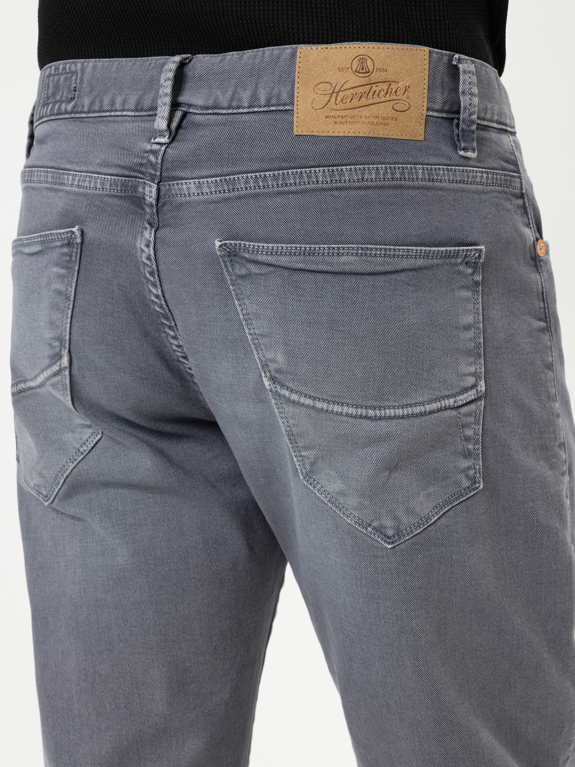 Nahaufnahme einer Person, die eine hellgraue Jeans trägt, von hinten gesehen, mit zwei Gesäßtaschen und einem braunen Herrlicher-Markenetikett am Bund.