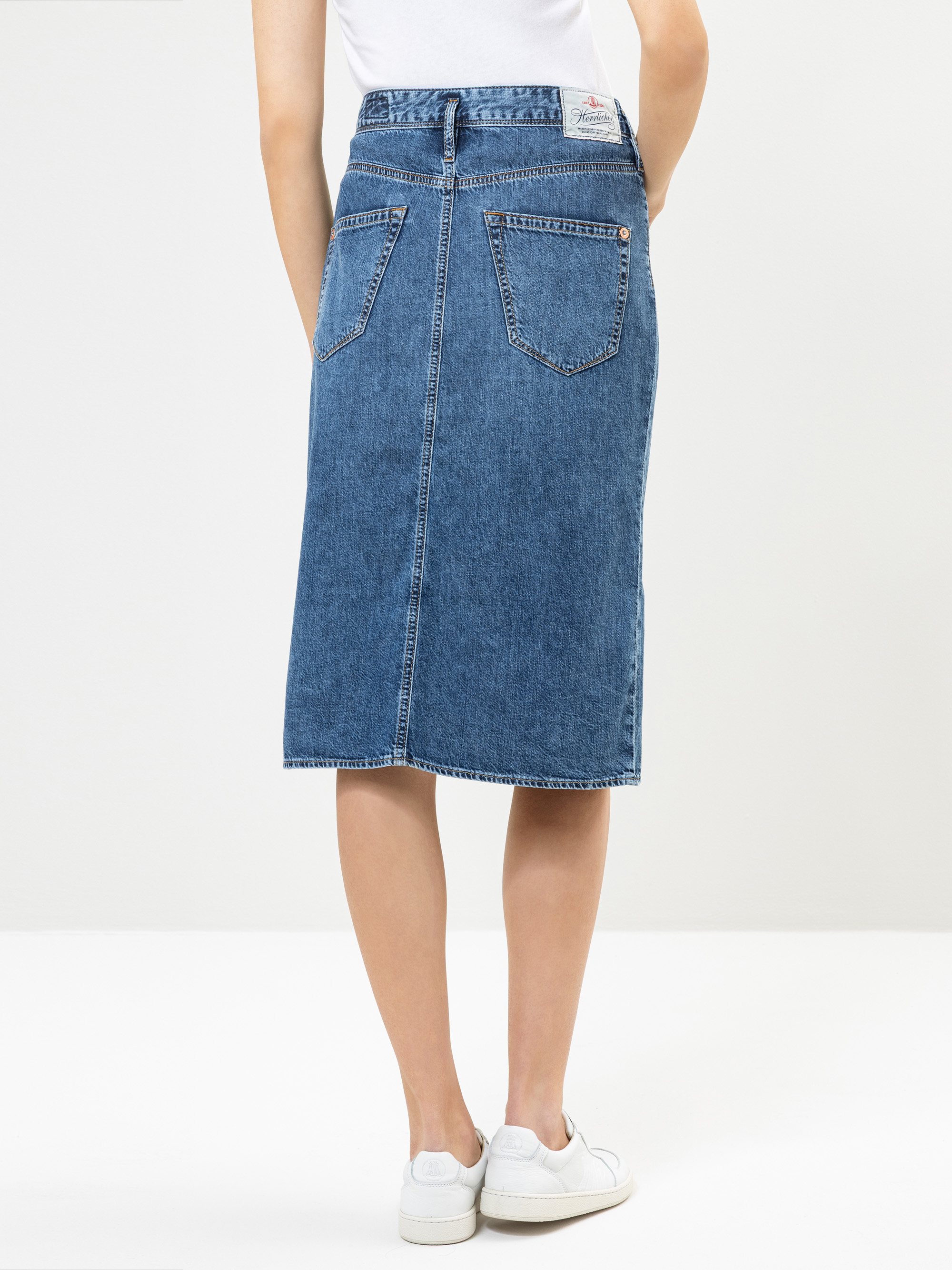 Eine Person, die einen blauen, knielangen Jeansrock, ein weißes Oberteil und weiße Turnschuhe trägt, wird von hinten gezeigt und steht vor einem schlichten, hellen Hintergrund.