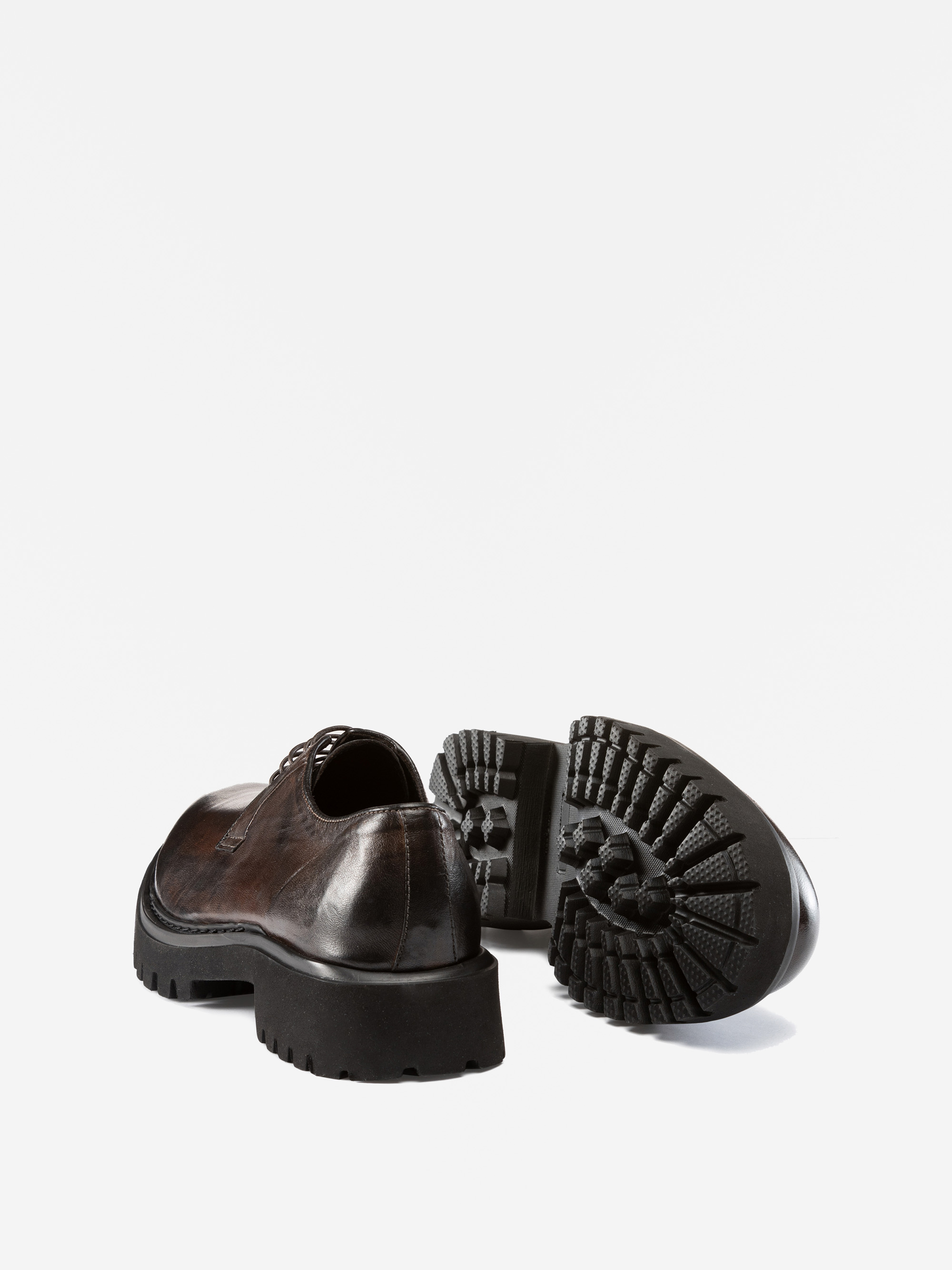 Abgebildet ist ein Paar dunkelbraune Lederschnürschuhe, von denen einer aufrecht steht und einer auf der Seite liegt, um die dicke, strukturierte schwarze Gummisohle vor einem schlichten weißen Hintergrund zu zeigen.