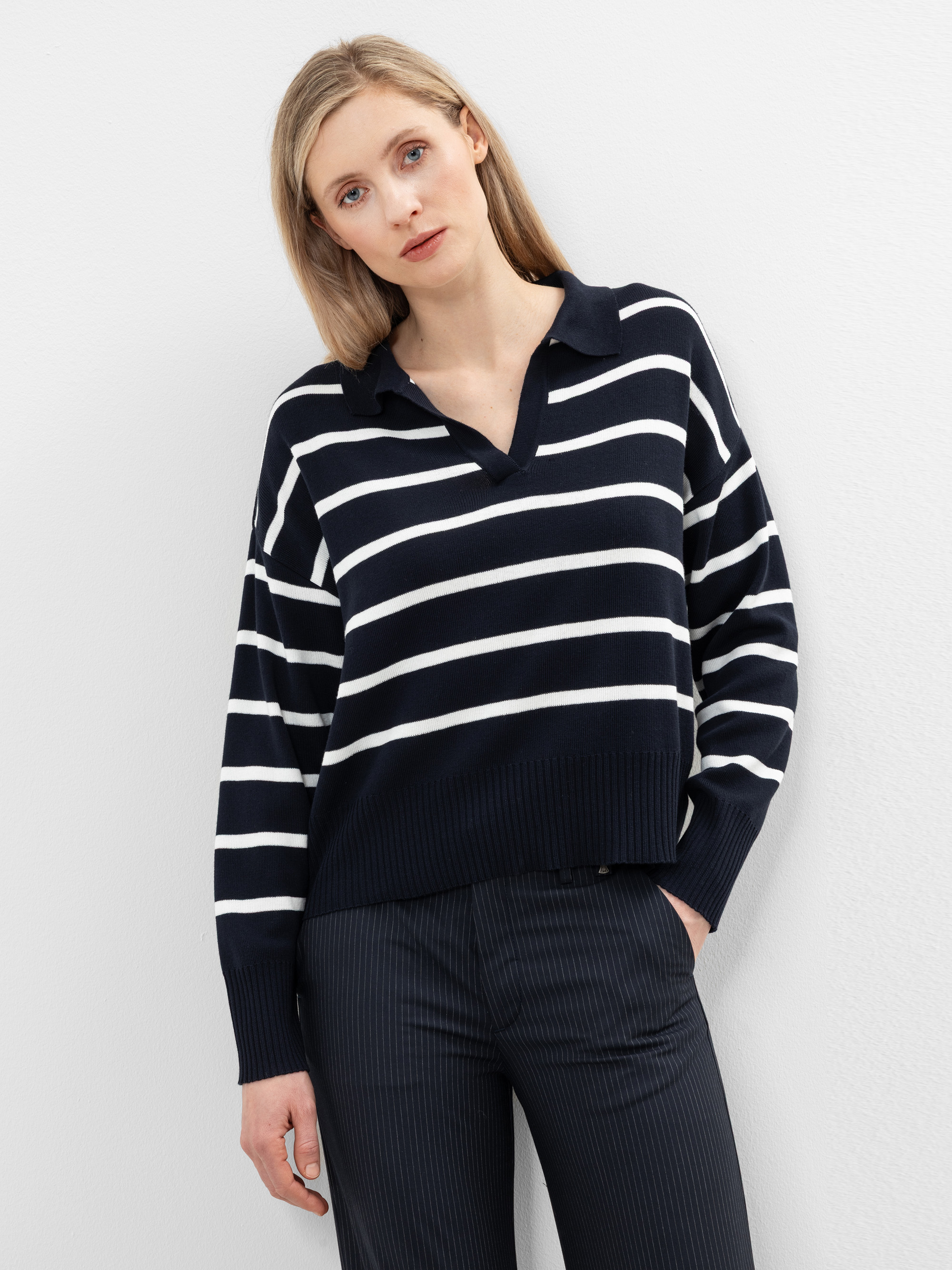 Eine Frau mit glattem blondem Haar trägt einen marineblauen Pullover mit weißen Querstreifen und eine dunkle Hose mit Nadelstreifen vor einem schlichten weißen Hintergrund.
