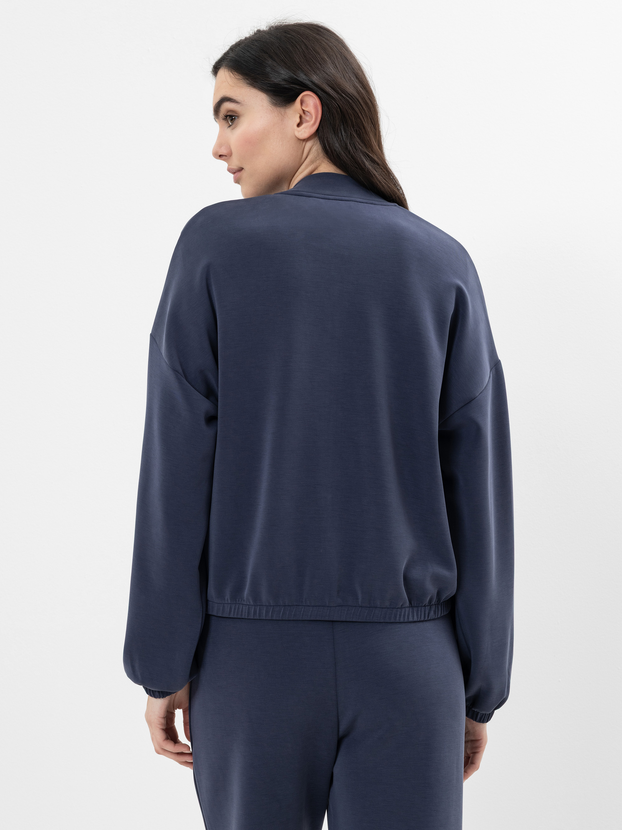 Eine Frau mit langen braunen Haaren steht mit dem Gesicht zur Seite und schaut über ihre Schulter. Sie trägt ein locker sitzendes, dunkelblaues Sweatshirt und eine passende Hose. Der Hintergrund ist einfarbig weiß.