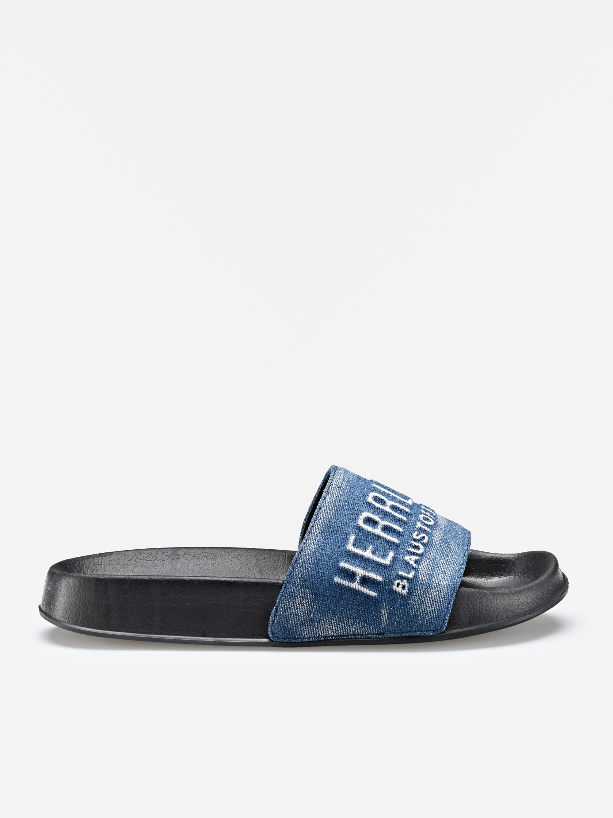 Eine einzelne schwarze Slide-Sandale mit einem blauen Jeansriemen und weißem gesticktem Text. Die Sandale ist auf einem einfarbig weißen Hintergrund in einer seitlichen Profilansicht zu sehen.