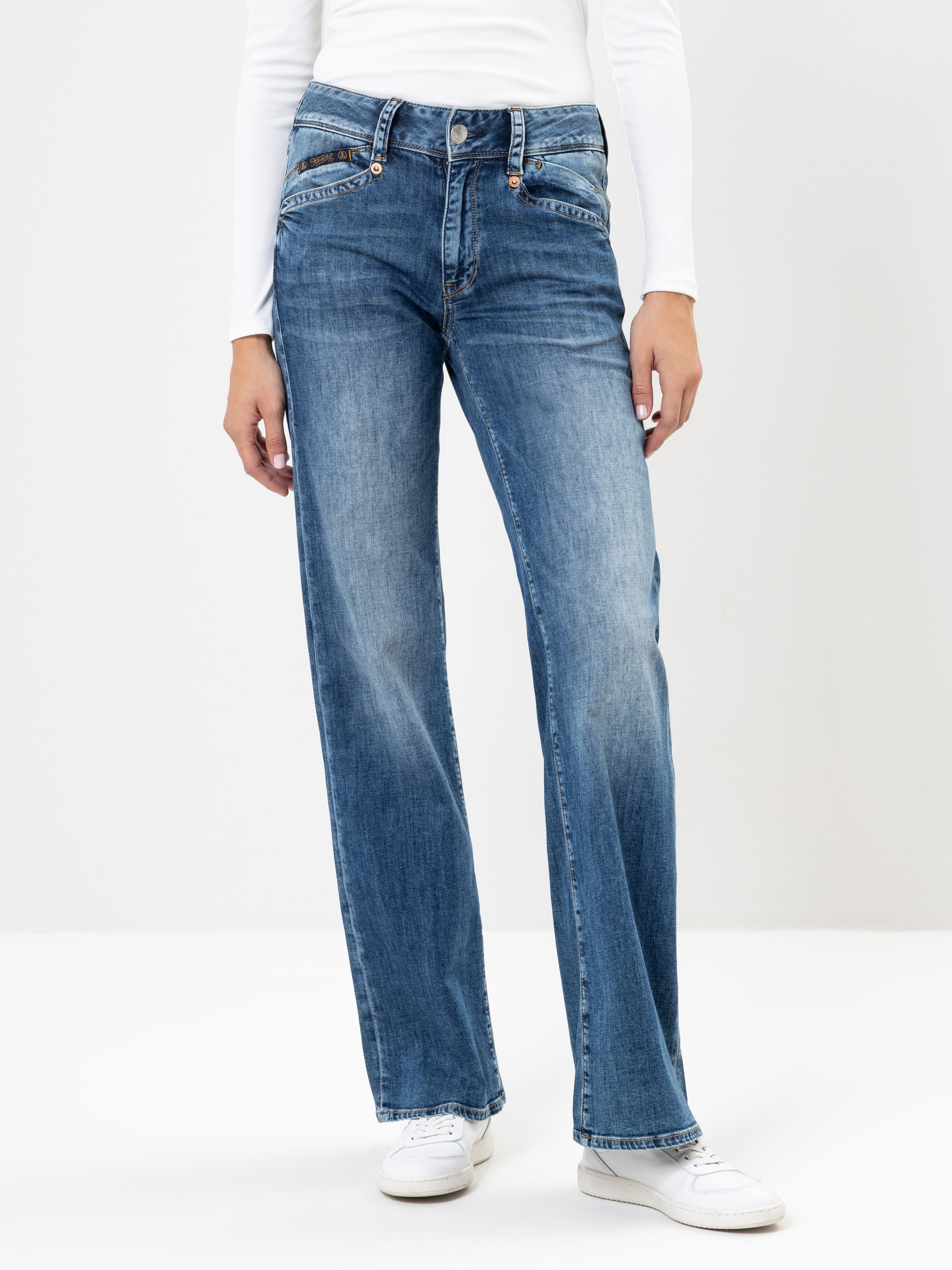 Eine Person, die blaue Jeans mit geradem Bein, ein weißes langärmeliges Oberteil und weiße Turnschuhe trägt, steht vor einem schlichten hellen Hintergrund.