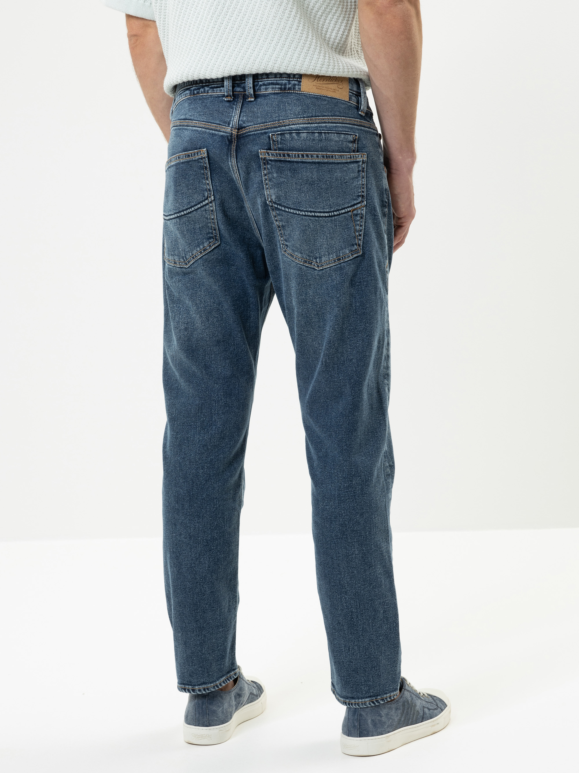 Eine Person, die blaue Jeans und helle Turnschuhe trägt, steht mit dem Rücken zur Kamera vor einem schlichten weißen Hintergrund. Die Jeans hat zwei Gesäßtaschen und einen hellbraunen Aufnäher am Bund.