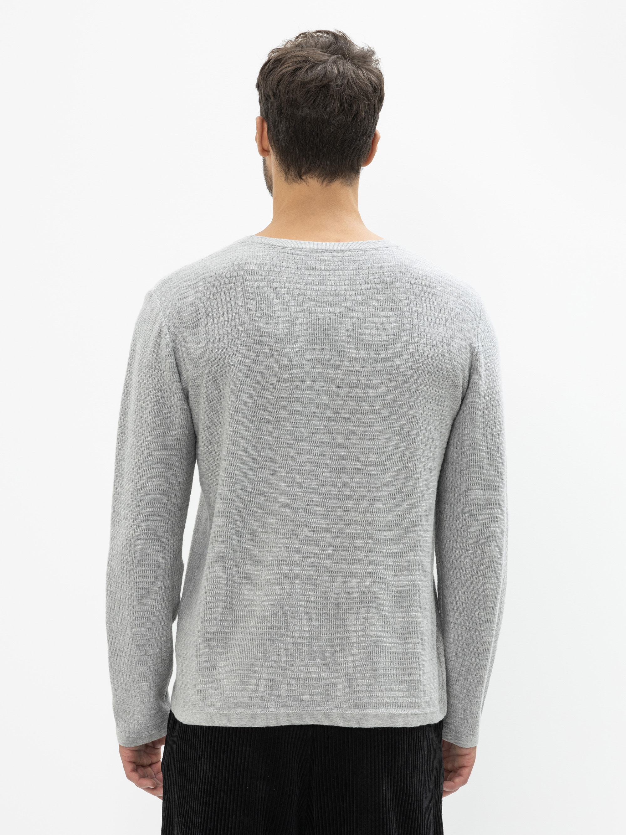 Ein Mann steht mit dem Rücken zur Kamera, trägt einen hellgrauen langärmeligen Pullover und eine dunkle Hose vor einem schlichten weißen Hintergrund.