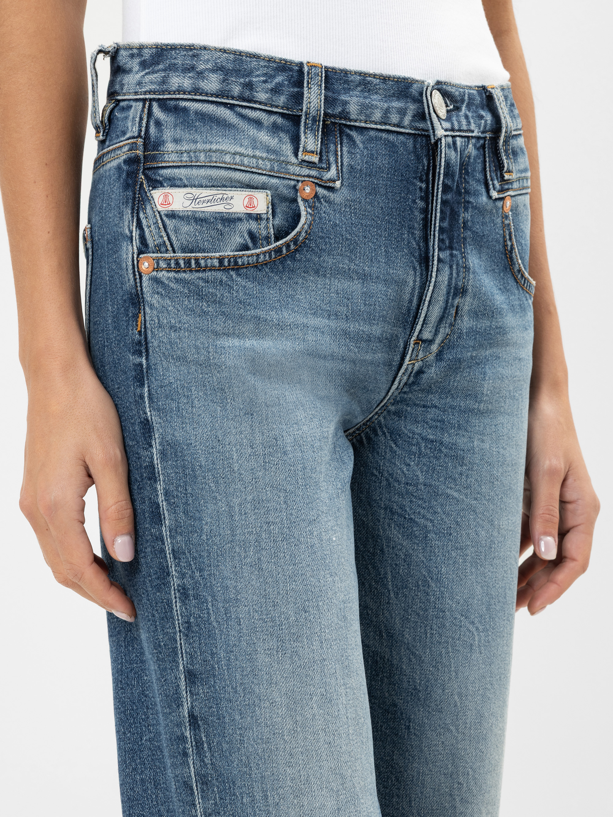 Nahaufnahme einer Person, die eine hoch taillierte Bluejeans und ein weißes Oberteil trägt und die Hände entspannt an der Seite hält. Die Jeans hat sichtbare Nähte, Kupfernieten und einen kleinen Aufnäher auf der Münztasche.
