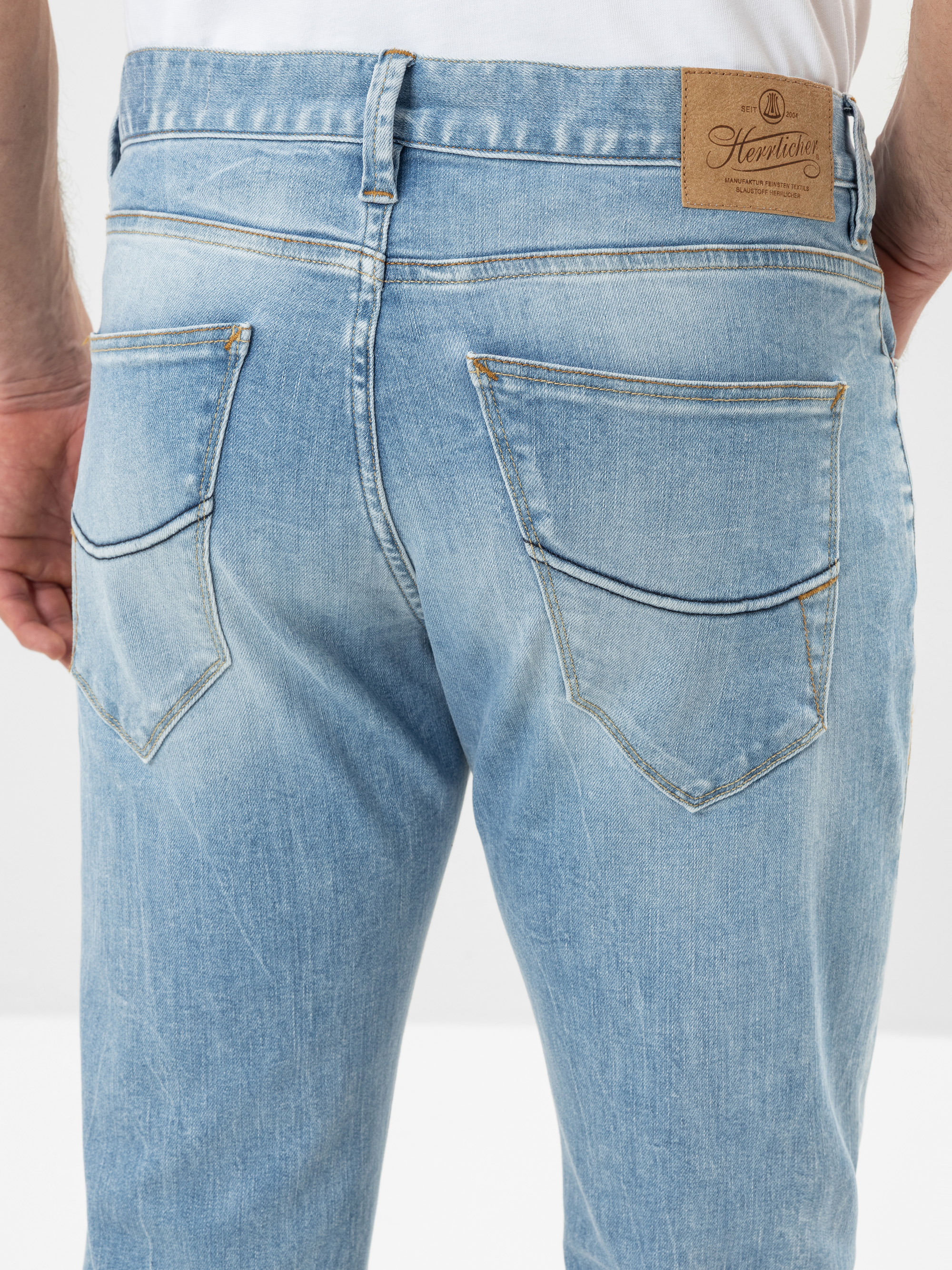 Nahaufnahme des Rückens einer Person, die eine hellblaue Denim-Jeans mit sichtbaren Taschen und Nähten trägt. Auf dem Bund ist ein hellbrauner Lederaufnäher mit einem Logo aufgenäht. Die Person steht vor einem einfarbigen Hintergrund.