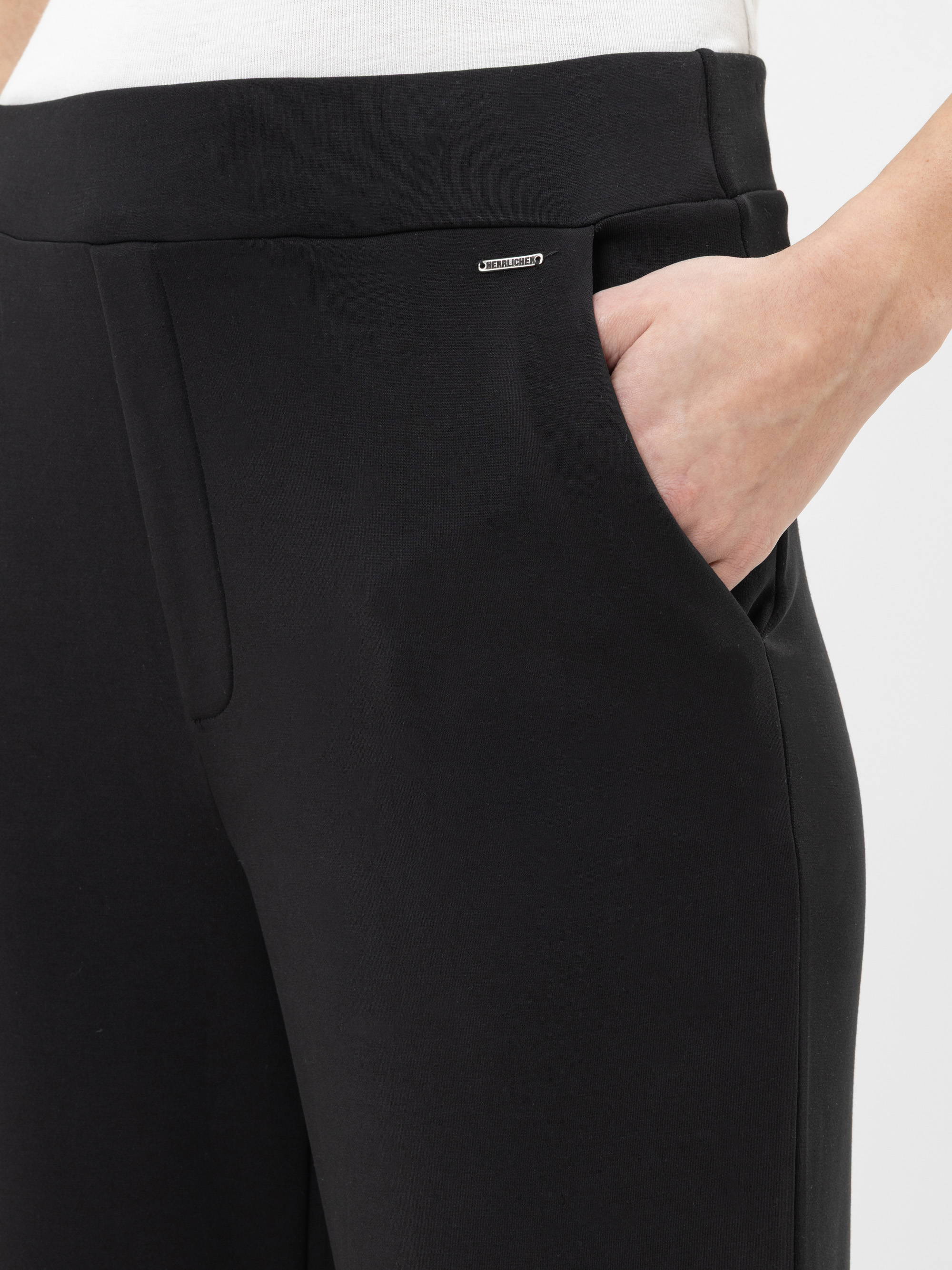 Nahaufnahme einer Person in einer schwarzen Hose mit einer Hand in der rechten Tasche. Die Hose hat eine glatte Struktur und ein kleines metallisches Markenetikett in der Nähe des Hosenbundes. Die Person trägt außerdem ein weißes Oberteil.