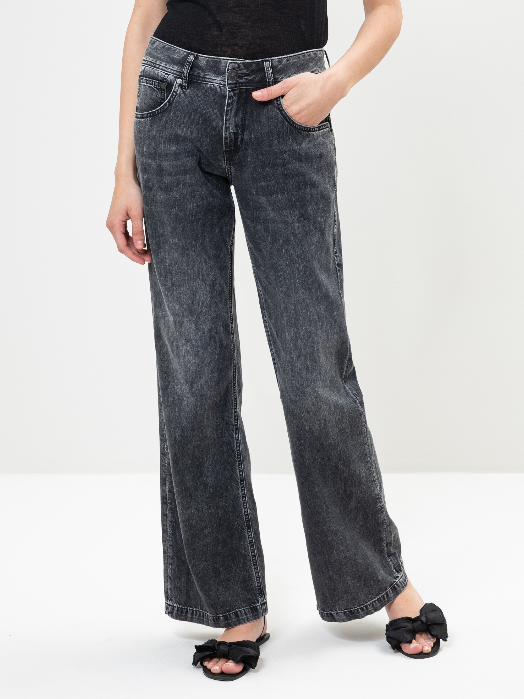 Eine Person, die eine schwarze verwaschene Jeans mit hoher Taille und weitem Bein, ein schwarzes Oberteil und schwarze offene Sandalen mit Schleifen trägt, steht vor einem schlichten weißen Hintergrund.