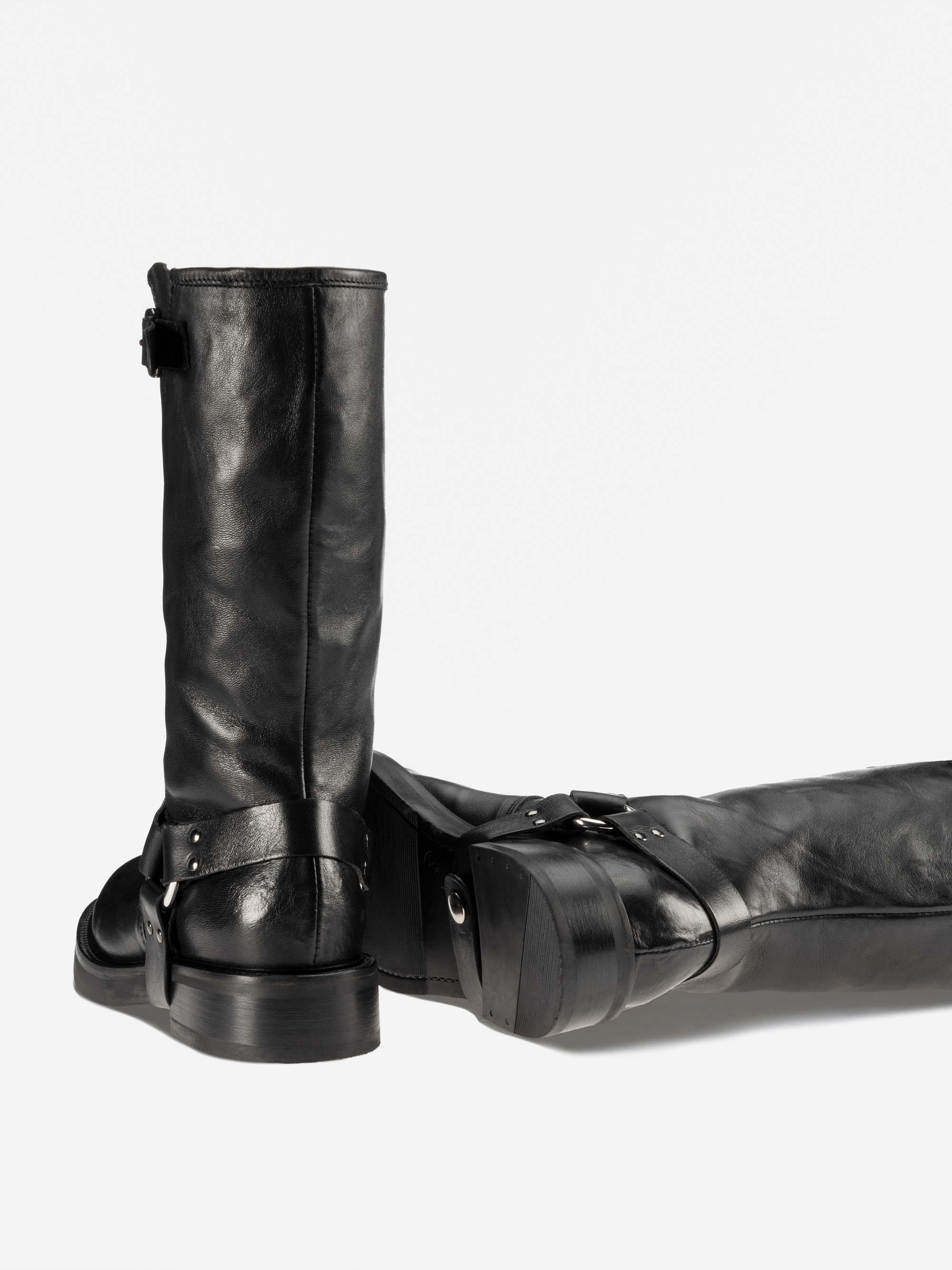 Ein Paar schwarzer Lederstiefel, einer aufrecht stehend, der andere auf der Seite liegend, beide mit einem Gurtband und einem niedrigen Blockabsatz auf einem schlichten weißen Hintergrund.