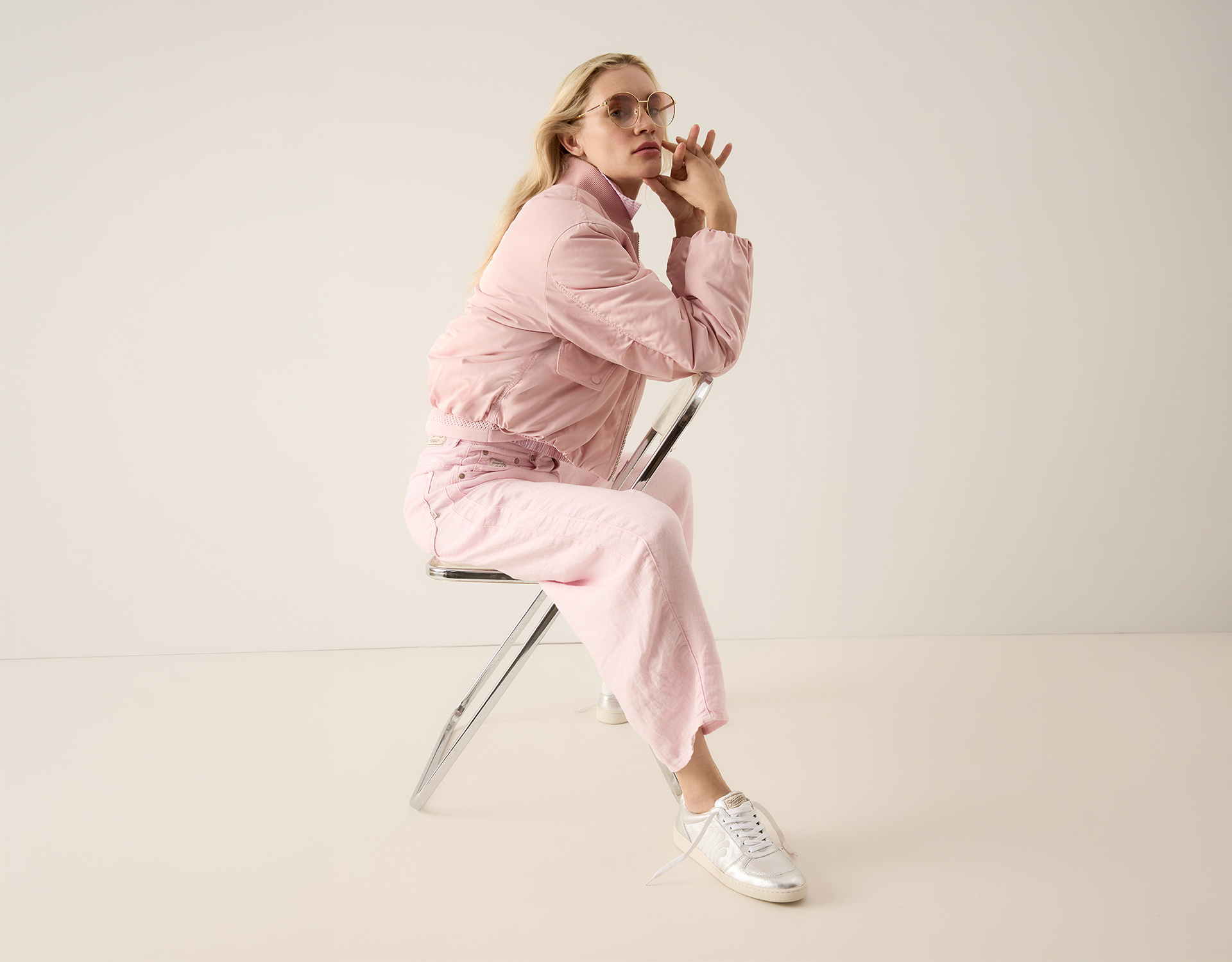 Person mit langen blonden Haaren, die eine hellrosa Jacke, eine rosa Hose und weiße Turnschuhe trägt und seitwärts auf einem modernen Metallstuhl vor einem schlichten hellen Hintergrund sitzt.