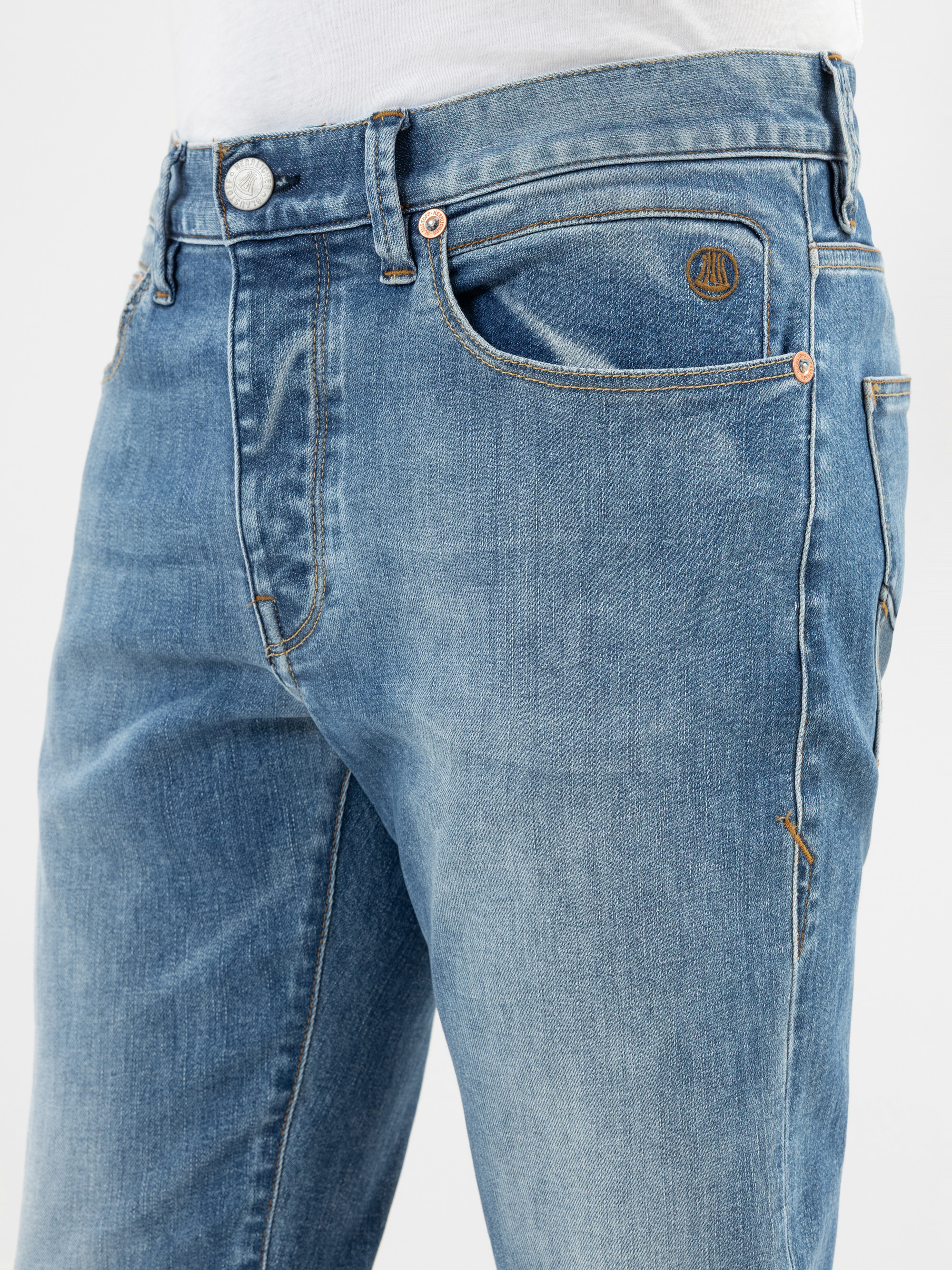 Nahaufnahme einer Person, die eine hellblaue Denim-Jeans und ein weißes Hemd trägt. Die Jeans hat Vordertaschen, kupferne Nieten und ein kleines gesticktes Logo über der rechten Tasche.