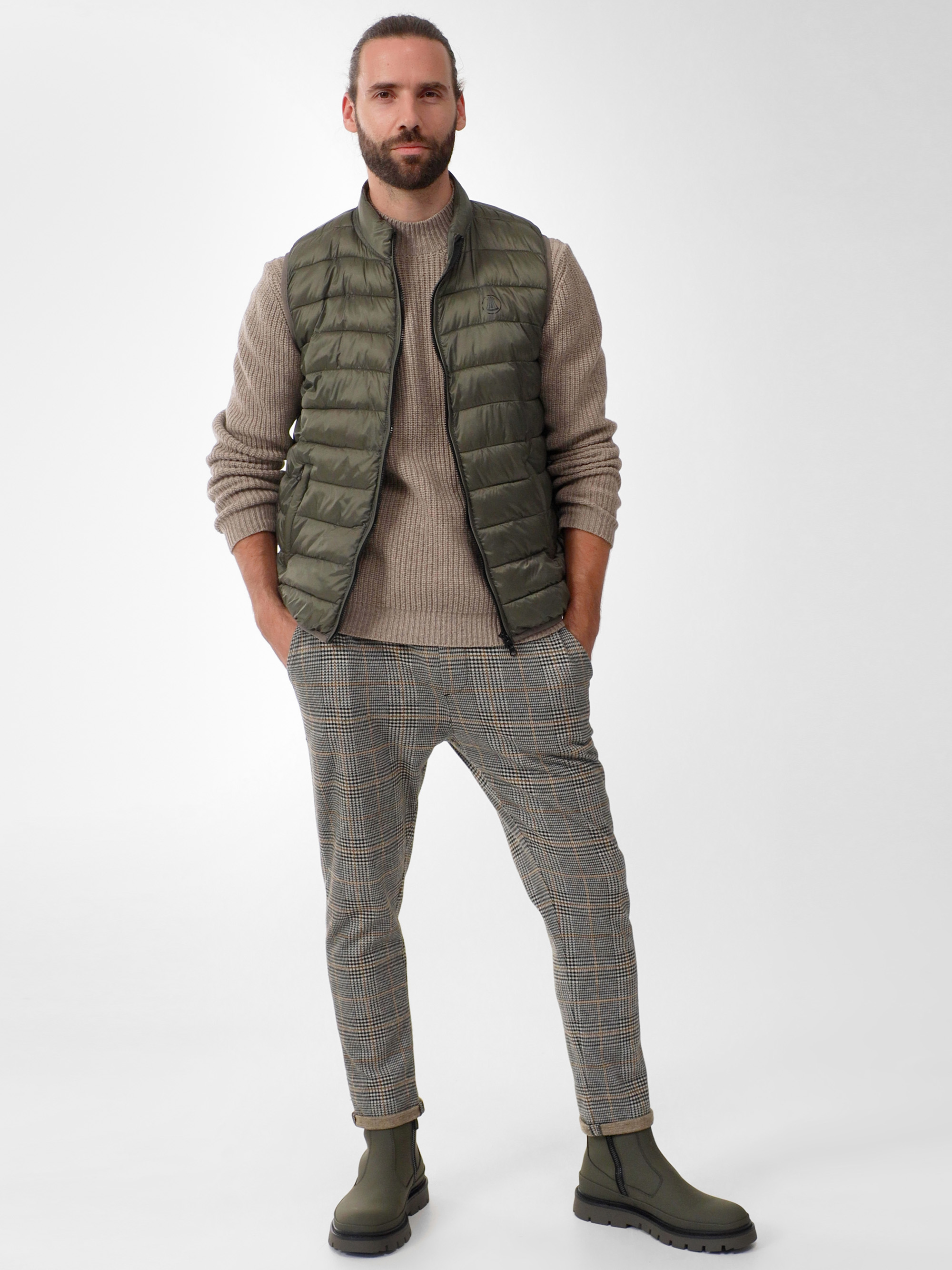 Ein Mann mit Bart steht vor einem schlichten Hintergrund und trägt eine olivgrüne Pufferweste, einen beigen Pullover, eine karierte Hose und grüne Stiefel, die Hände in den Taschen.