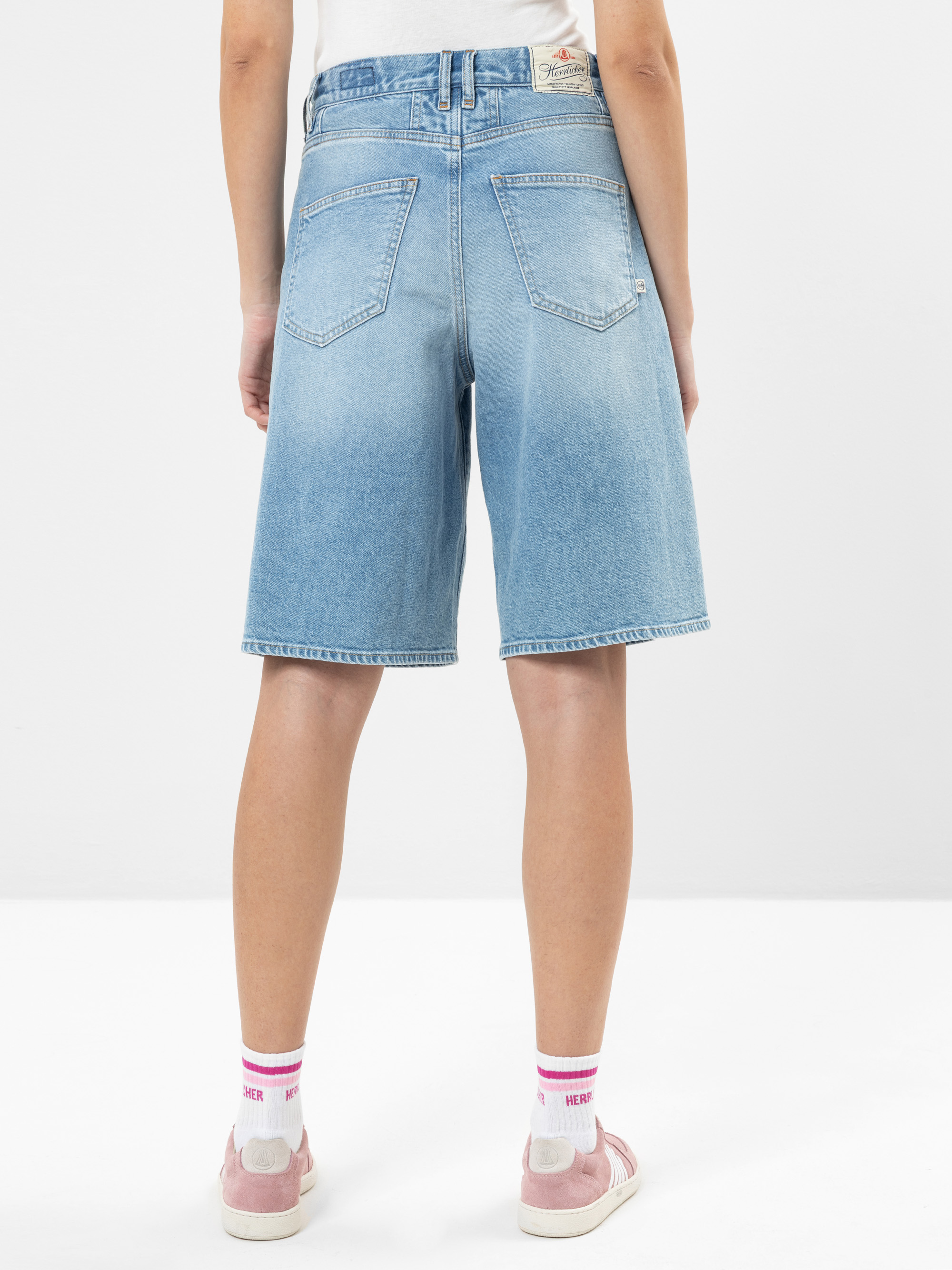 Eine Person steht mit dem Gesicht zur Wand, trägt hellblaue knielange Jeans-Shorts, ein weißes Oberteil, weiße und rosa Socken und rosa Turnschuhe vor einem schlichten weißen Hintergrund.