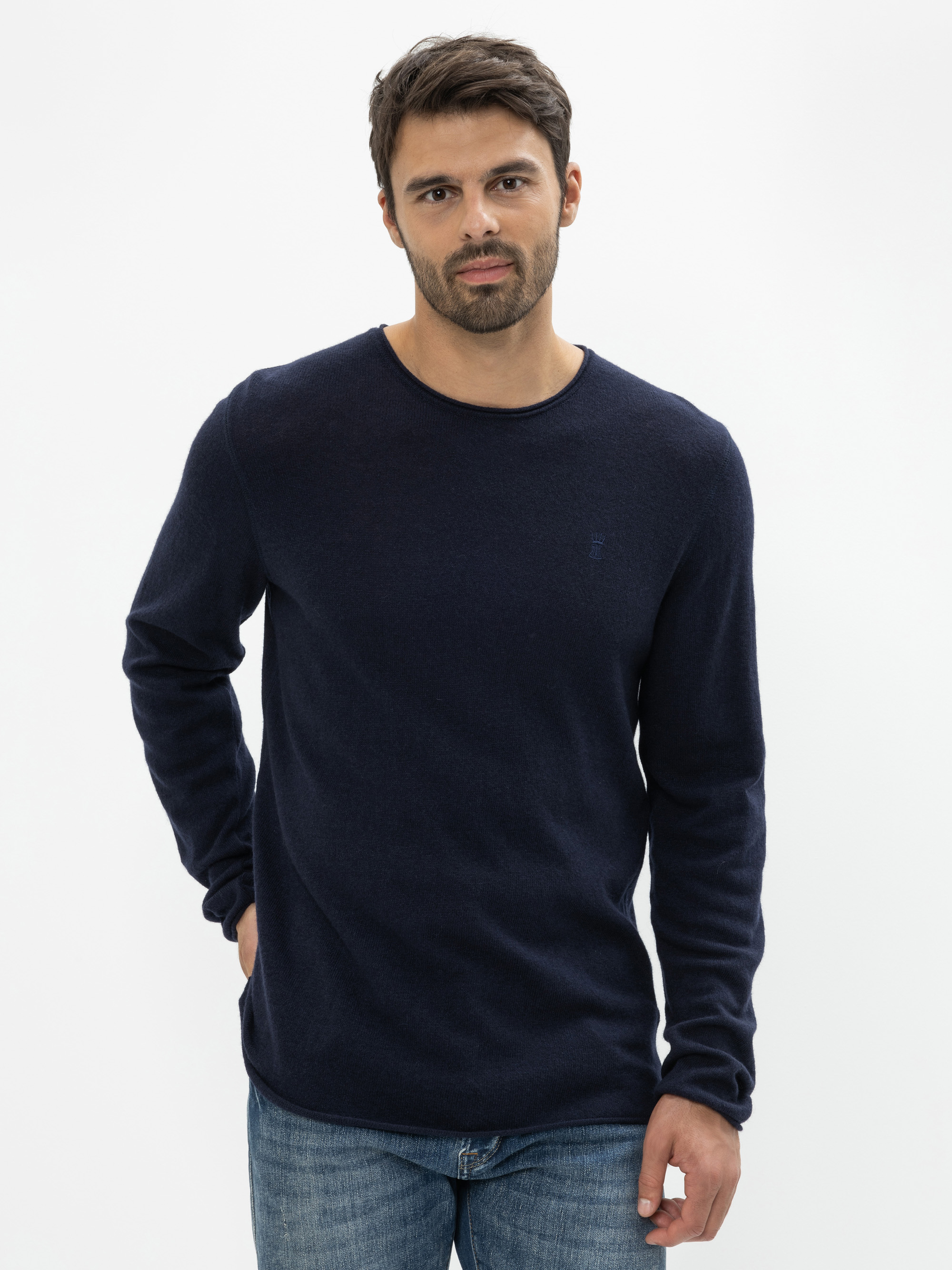 Ein Mann mit kurzen dunklen Haaren und einem Bart trägt einen marineblauen langärmeligen Pullover und blaue Jeans und steht mit neutralem Gesichtsausdruck vor einem schlichten weißen Hintergrund.