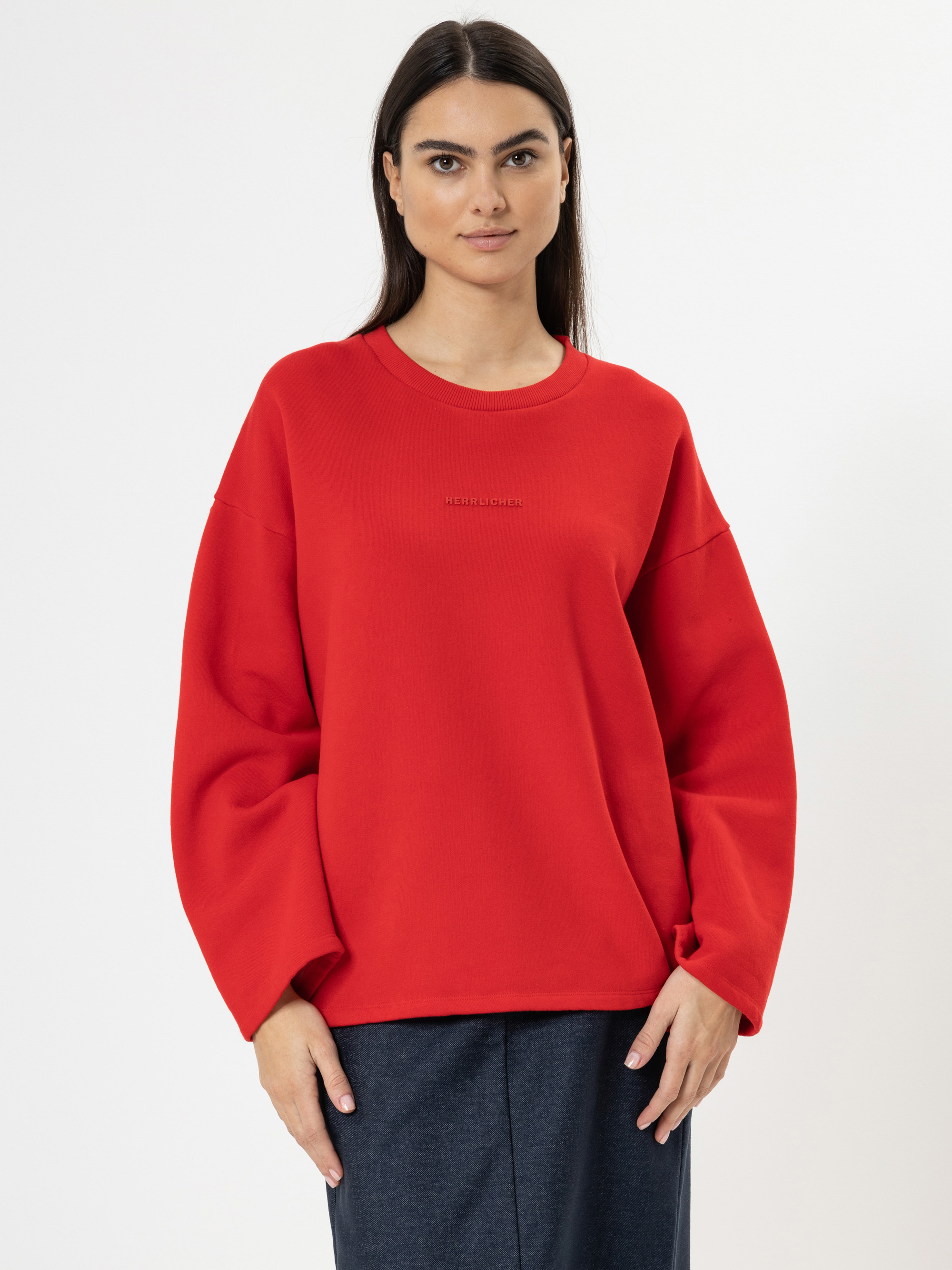 Eine Frau mit langen dunklen Haaren trägt ein leuchtend rotes Sweatshirt mit weiten Ärmeln und eine dunkle Hose und steht vor einem schlichten weißen Hintergrund. Sie blickt nach vorne und hat einen neutralen Gesichtsausdruck.