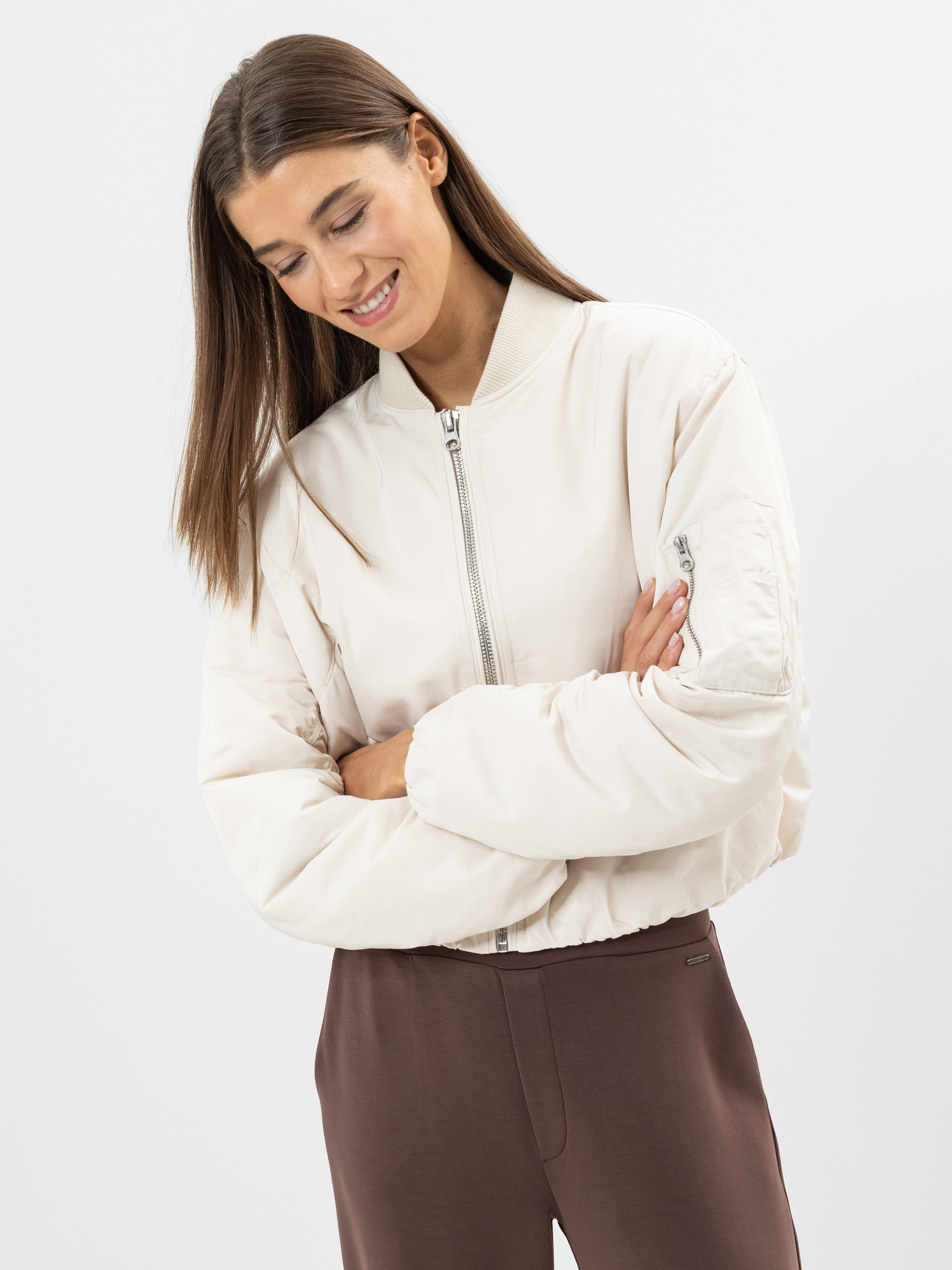 Eine Frau mit glattem braunem Haar trägt eine hellbeige Bomberjacke mit einem Frontreißverschluss und eine braune Hose. Sie lächelt und schaut mit verschränkten Armen vor einem schlichten weißen Hintergrund nach unten.