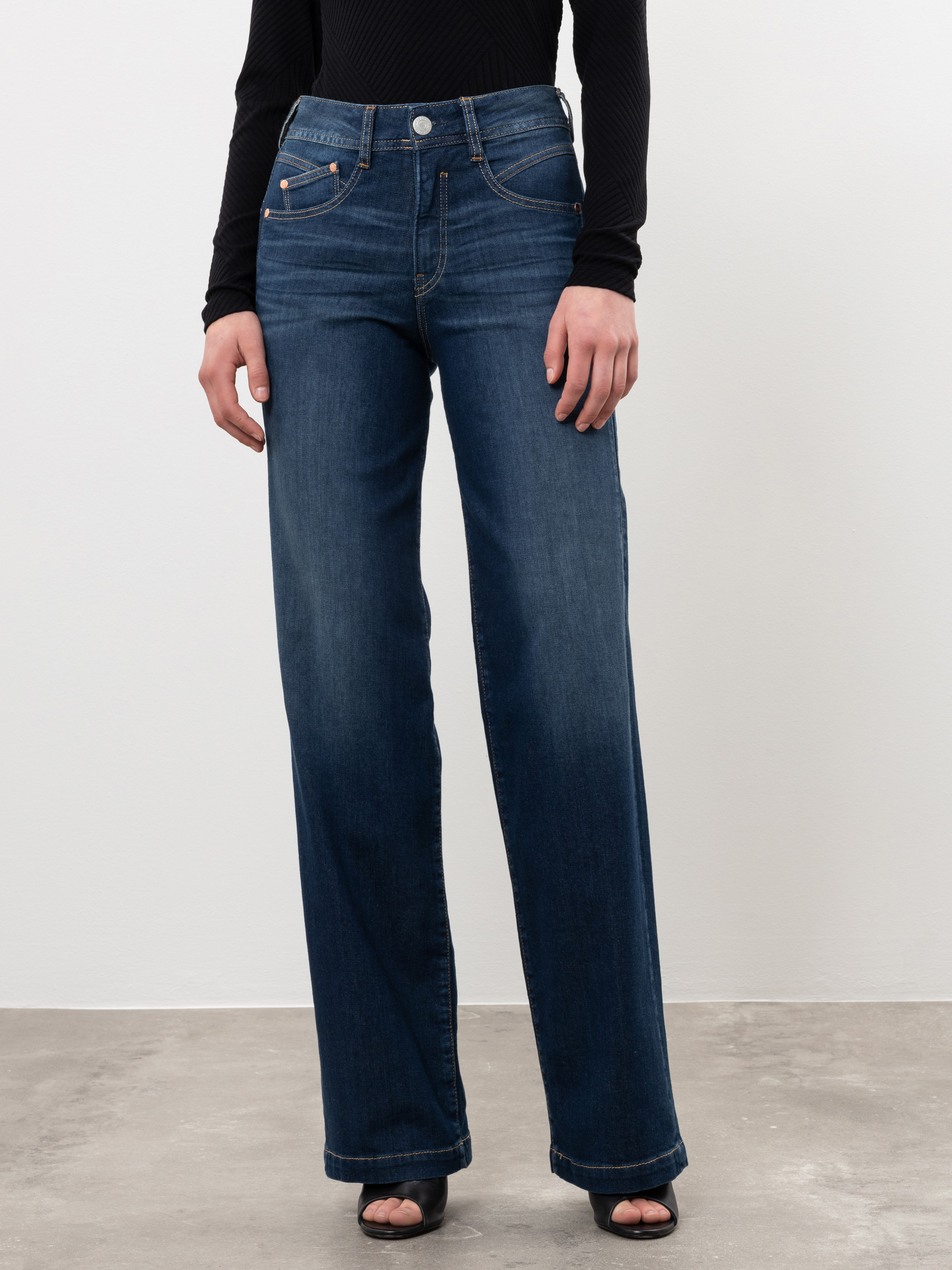 Eine Person steht vor einem unifarbenen Hintergrund und trägt eine hochgeschnittene, weit geschnittene Bluejeans und ein schwarzes, langärmeliges Oberteil; ihre Hände sind locker an der Seite und sie trägt schwarze, offene Schuhe.