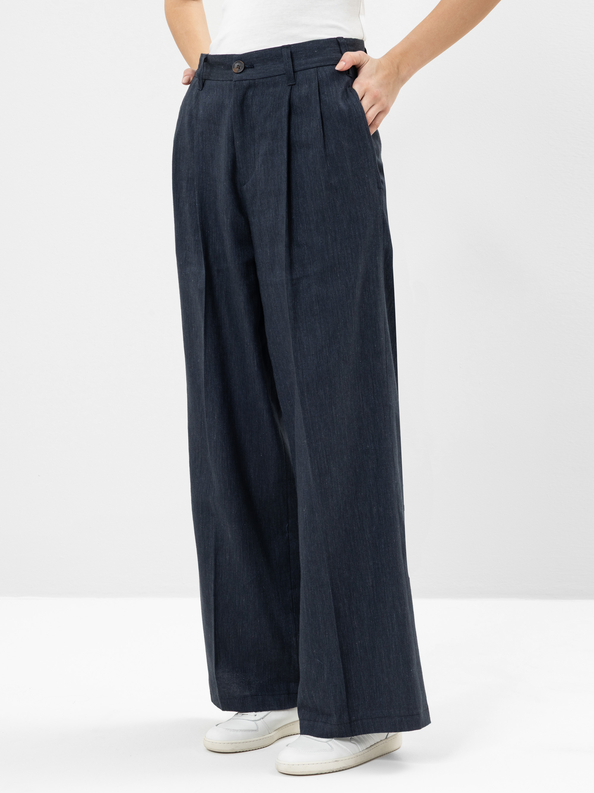 Eine Person, die eine dunkle Jeanshose mit hoher Taille und weitem Bein trägt, die vorne in Falten gelegt ist, gepaart mit einem weißen Oberteil und weißen Turnschuhen, steht vor einem schlichten weißen Hintergrund.