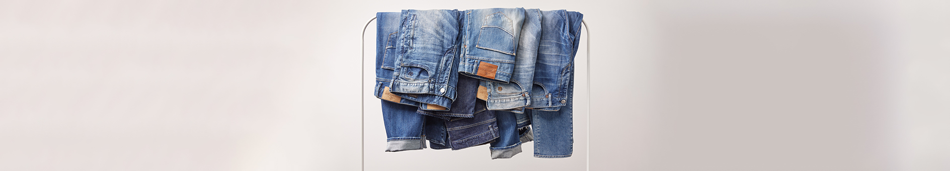 Mehrere Paar blaue Jeans sind vor einem hellen, neutralen Hintergrund über eine horizontale Stange drapiert. Die Jeans variieren in den Blautönen und weisen Anzeichen von Abnutzung und Verblassen auf.
