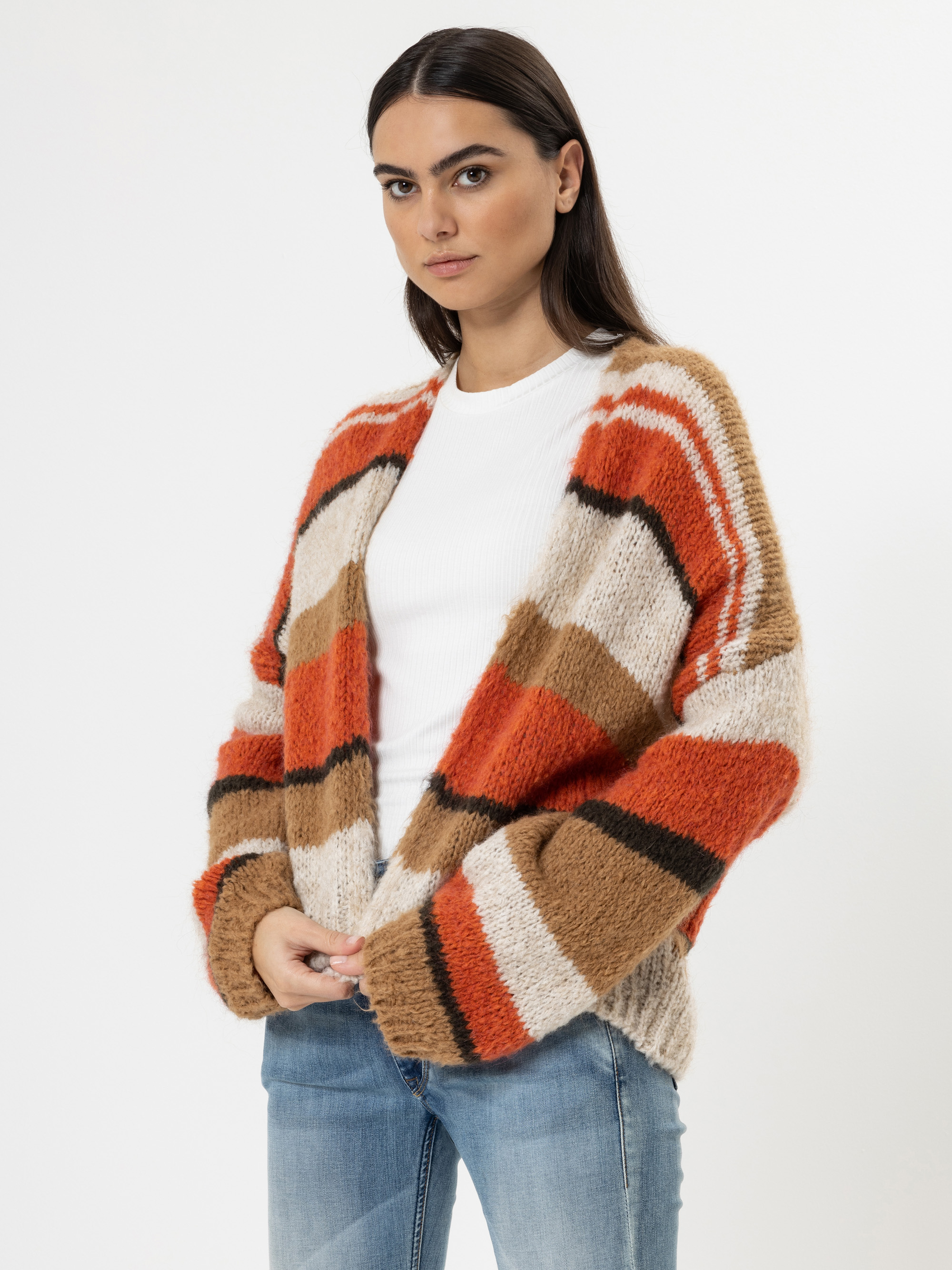 Eine Frau mit langen dunklen Haaren, die ein weißes geripptes Hemd, eine gestreifte Strickjacke in Orange-, Beige-, Braun- und Cremetönen und hellblaue Jeans trägt, steht vor einem schlichten weißen Hintergrund.