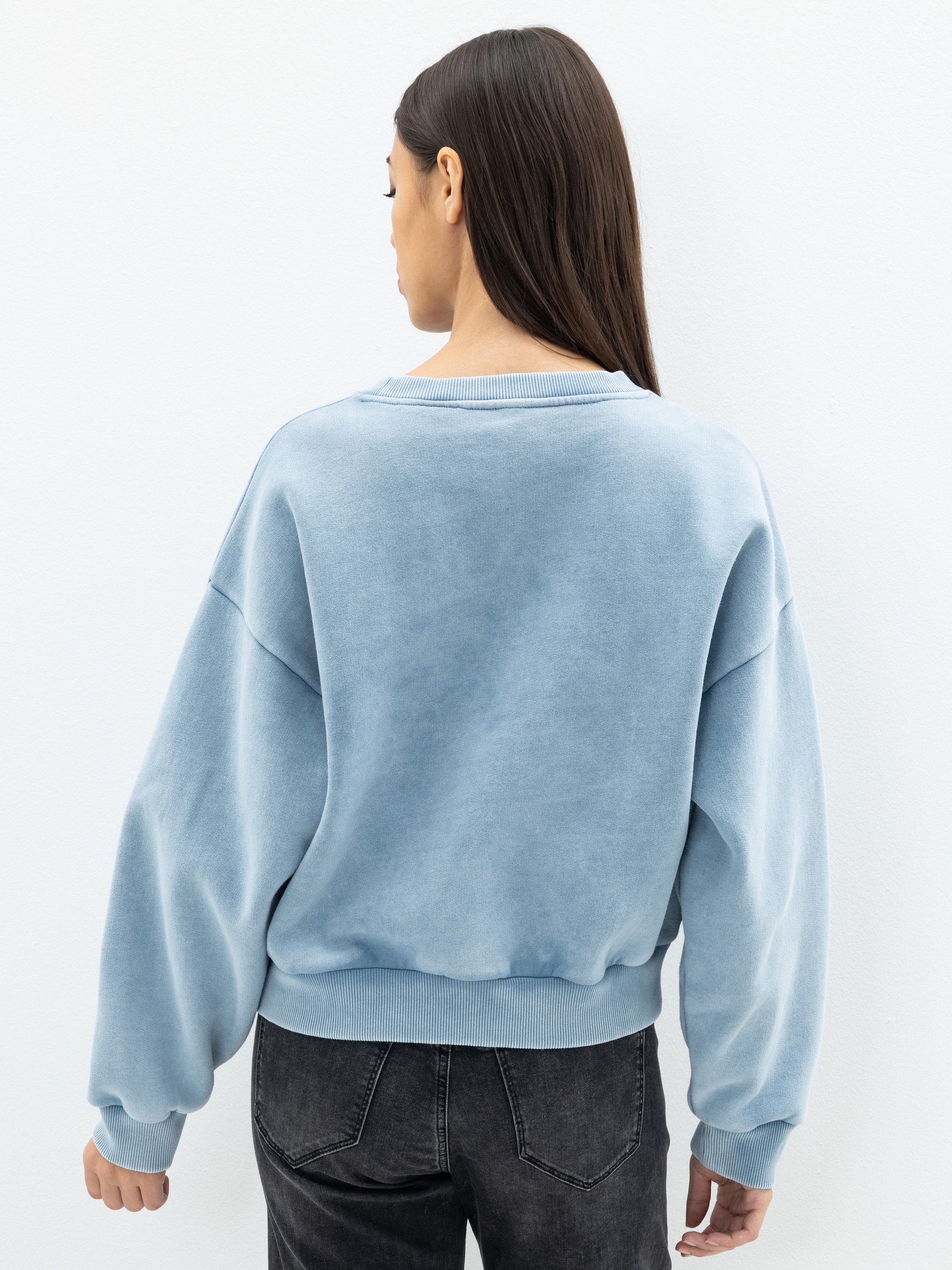 Eine Frau mit langen dunklen Haaren, die ein hellblaues Sweatshirt und schwarze Jeans trägt, steht mit dem Gesicht zur Kamera vor einem schlichten weißen Hintergrund.