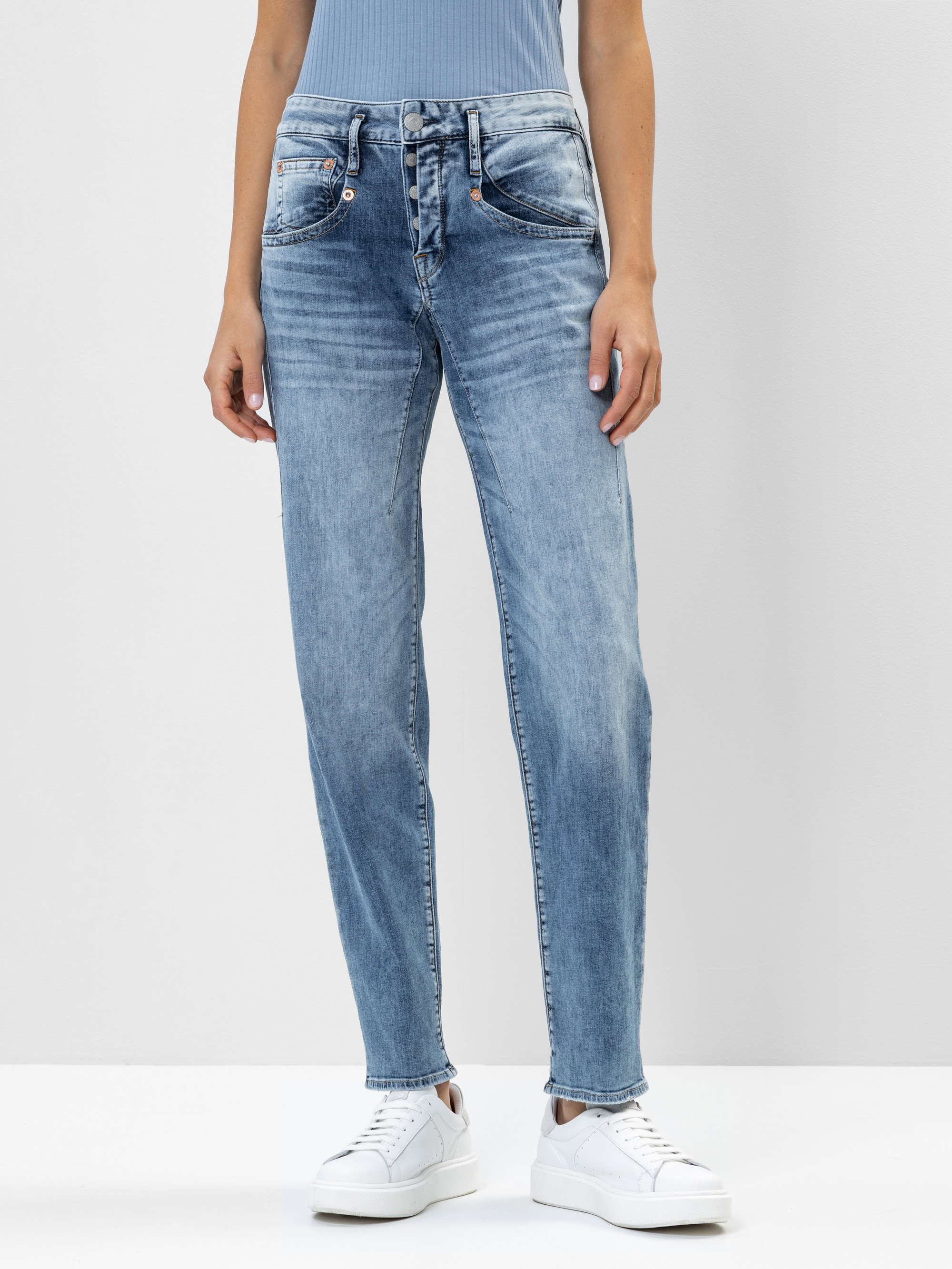 Eine Person trägt eine hellblaue, hoch taillierte Jeans, ein blaues, geripptes Oberteil und weiße Turnschuhe vor einem schlichten, hellen Hintergrund. Die Jeans hat eine lockere Passform und verblasste Details.