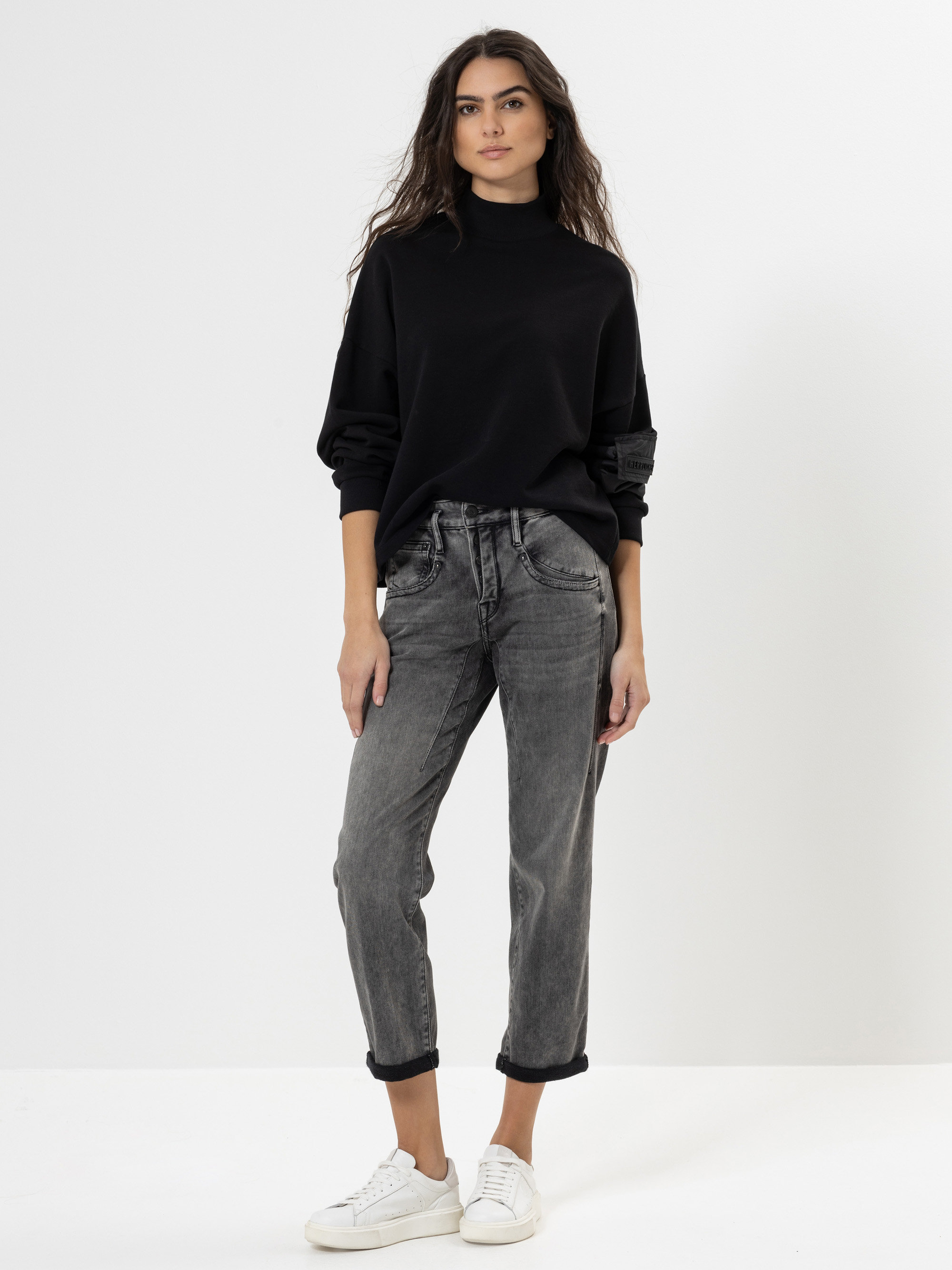 Eine Frau steht vor einem weißen Hintergrund und trägt einen schwarzen Rollkragenpullover, graue Jeans mit Bündchen und weiße Turnschuhe. Ihre Hände sind entspannt, und sie hat langes, gewelltes braunes Haar.