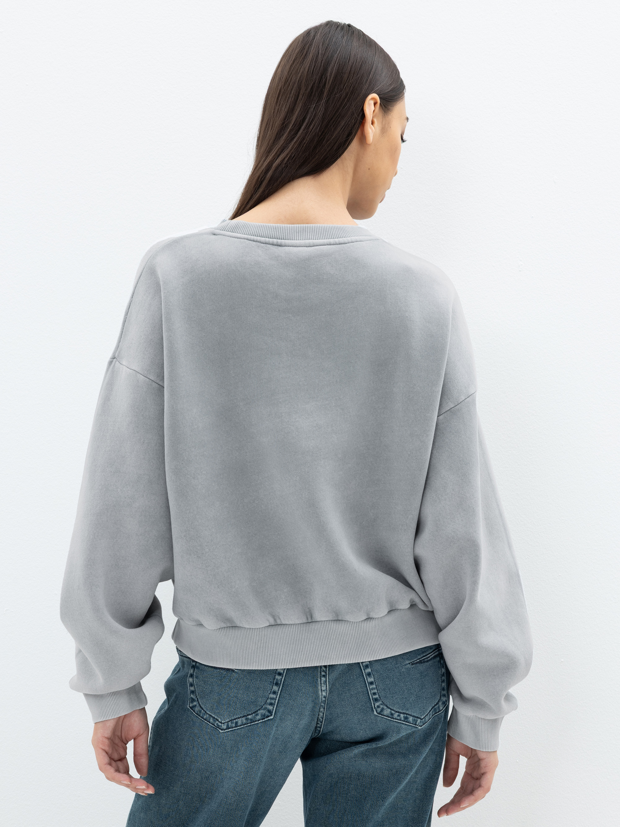 Eine Frau mit langen braunen Haaren, die ein locker sitzendes, hellgraues Sweatshirt und blaue Jeans trägt, steht mit dem Gesicht zur Kamera vor einem schlichten weißen Hintergrund.