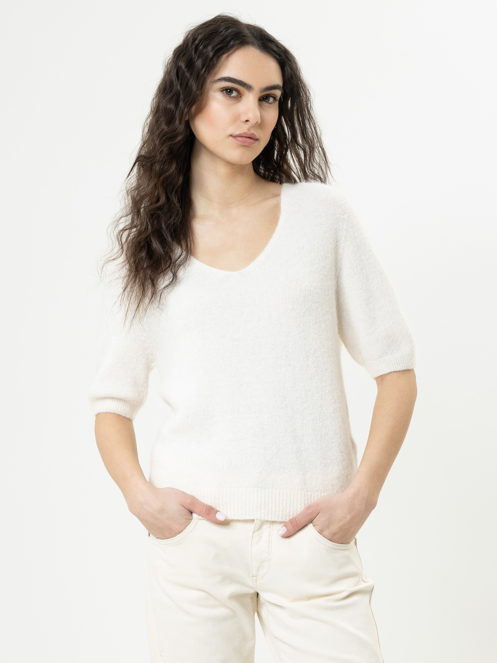 Eine Frau mit langen, gewellten dunklen Haaren trägt einen weichen, weißen, kurzärmeligen Pullover und eine helle Hose. Sie steht vor einem schlichten weißen Hintergrund, hat die Hände in den Taschen und blickt selbstbewusst nach vorne.