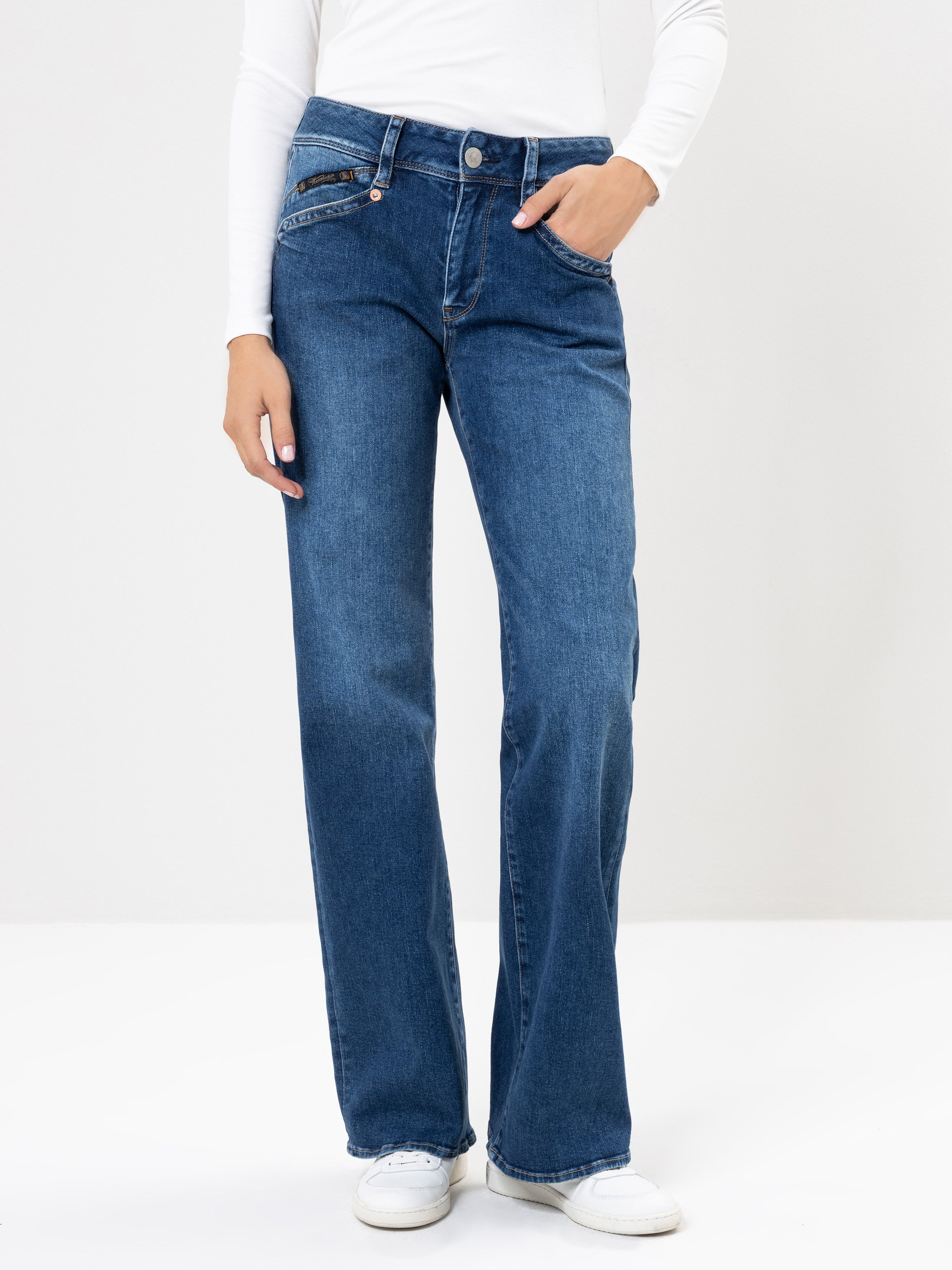 Person, die ein langärmeliges weißes Oberteil, eine blaue Jeans mit hohem Bund und Seitentaschen sowie weiße Turnschuhe trägt und vor einem schlichten weißen Hintergrund steht.