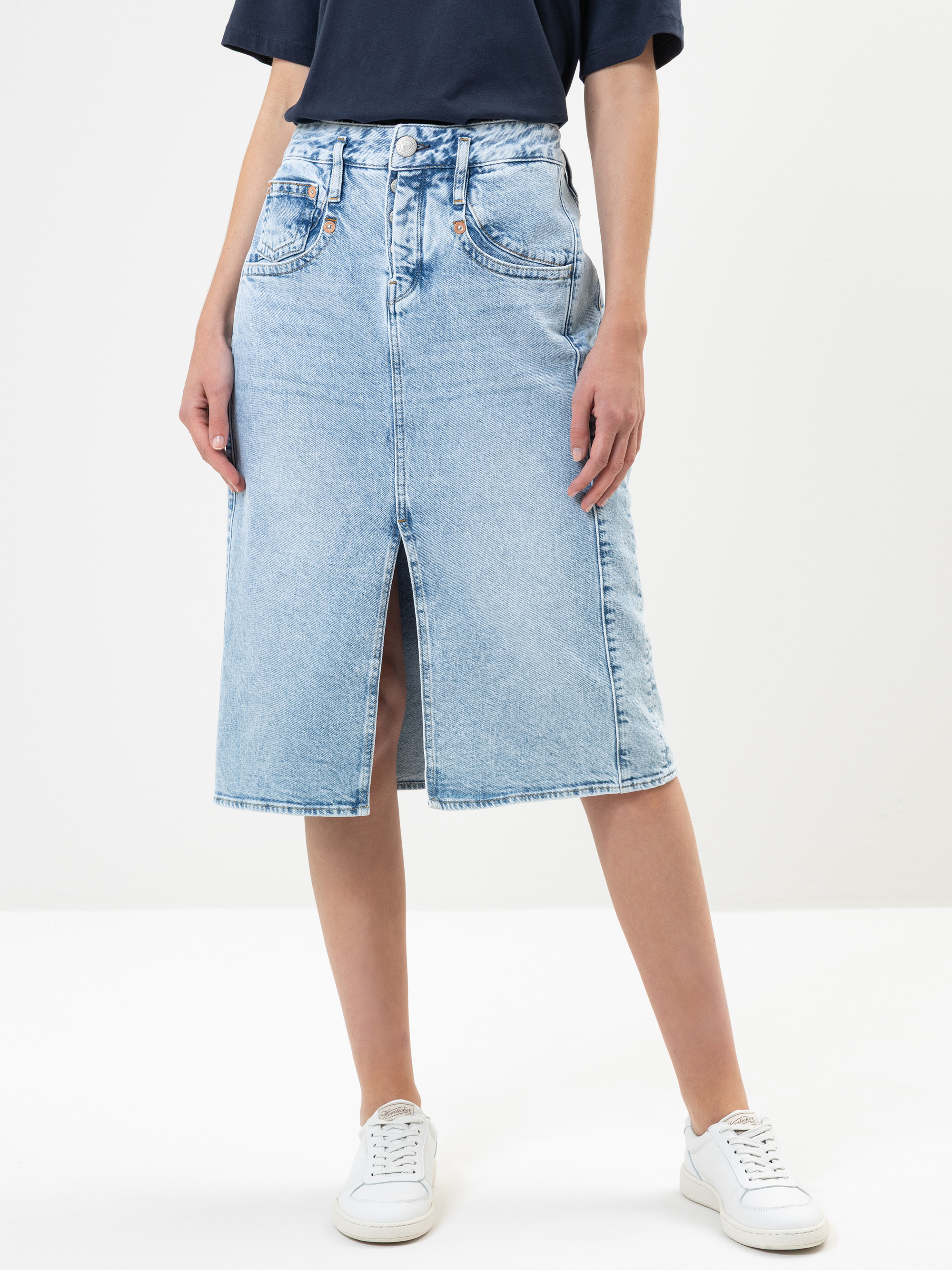 Eine Person, die einen knielangen, hellblauen Jeansrock mit einem Schlitz an der Vorderseite trägt, kombiniert mit einem marineblauen T-Shirt und weißen Turnschuhen, steht vor einem schlichten hellen Hintergrund.
